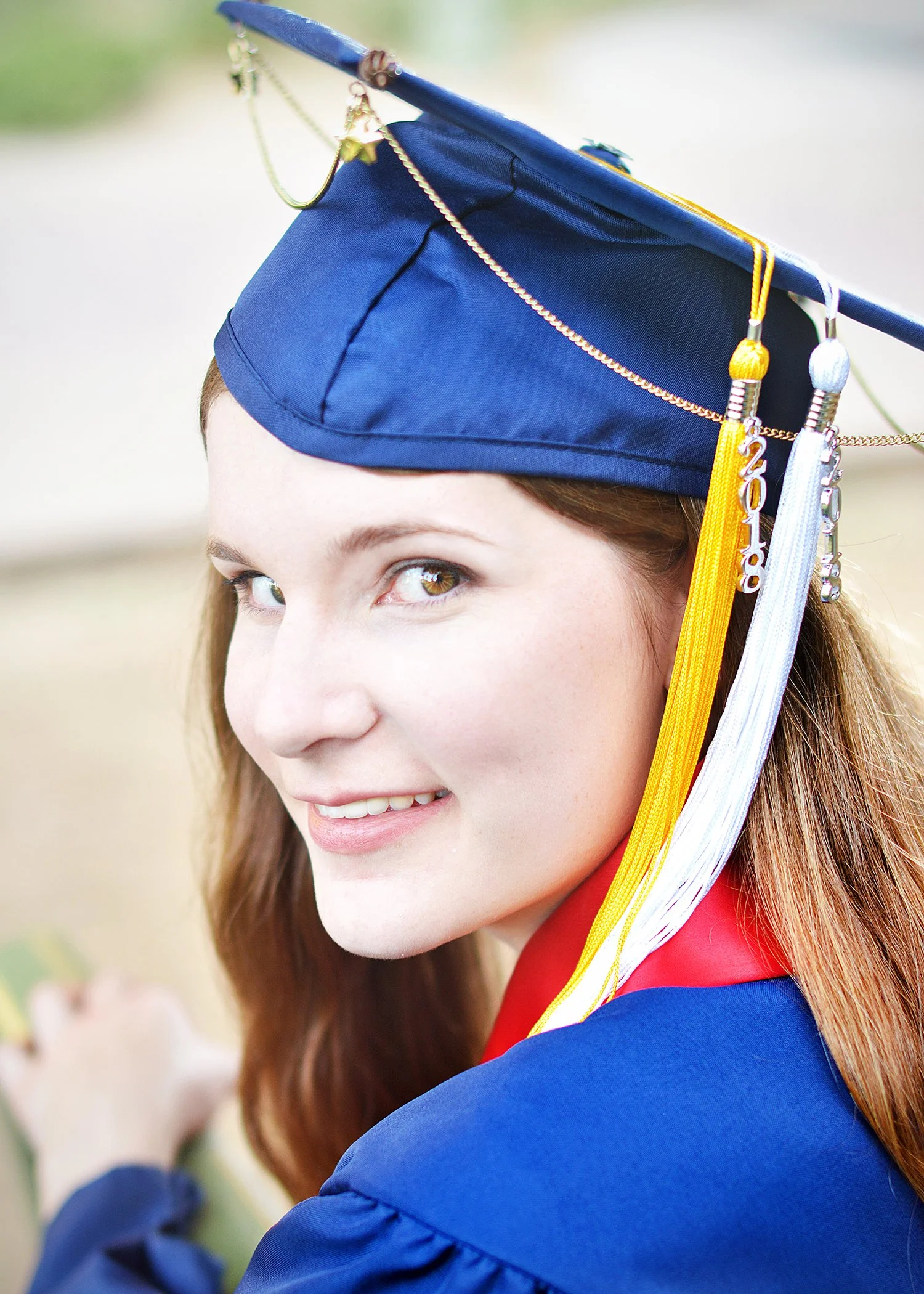 Close-up of a young woman with long brown hair wearing a blue graduation cap from University Arizona and gown, smiling with green eyes, outdoors, celebration during a senior photo shoot in Tucson, Arizona
