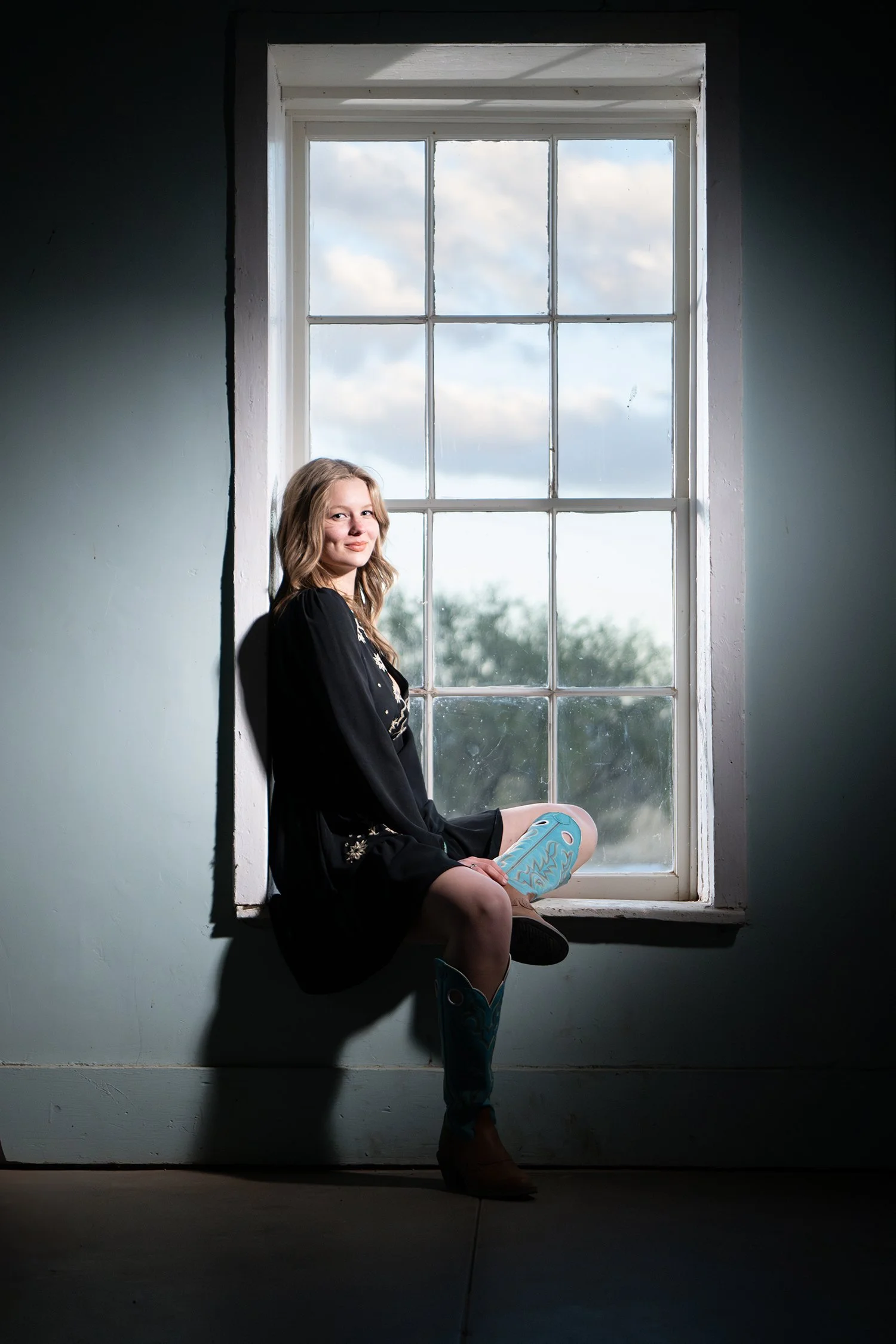 A young woman with light brown hair sitting on a windowsill, wearing a black dress and turquoise cowboy boots with a decorative design, looking at the camera with a slight smile. The window behind her shows a cloudy sky with trees in Tucson