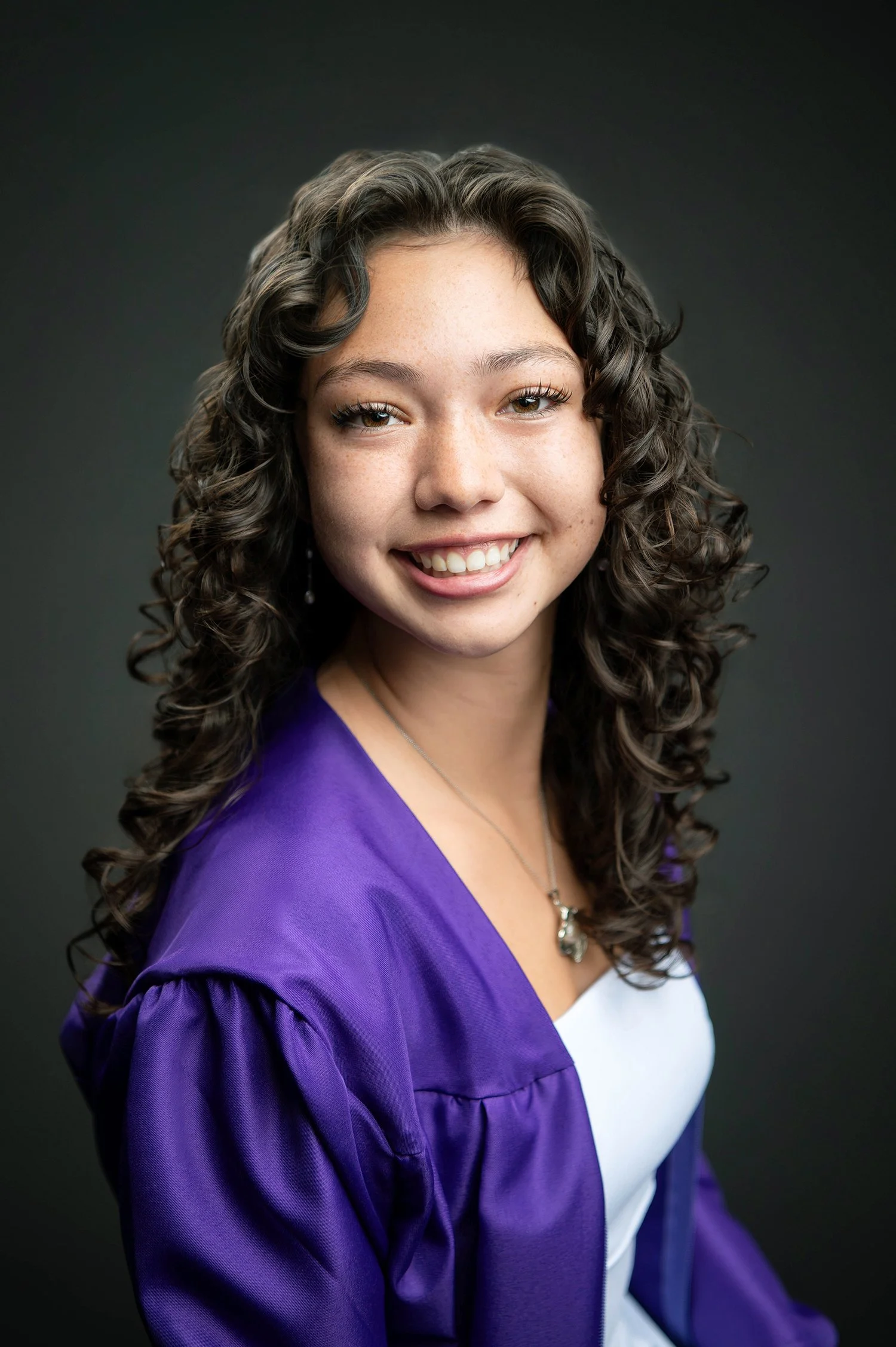 Portrait of a young woman with curly brown hair, wearing a purple jacket over a white top, smiling against a dark background.