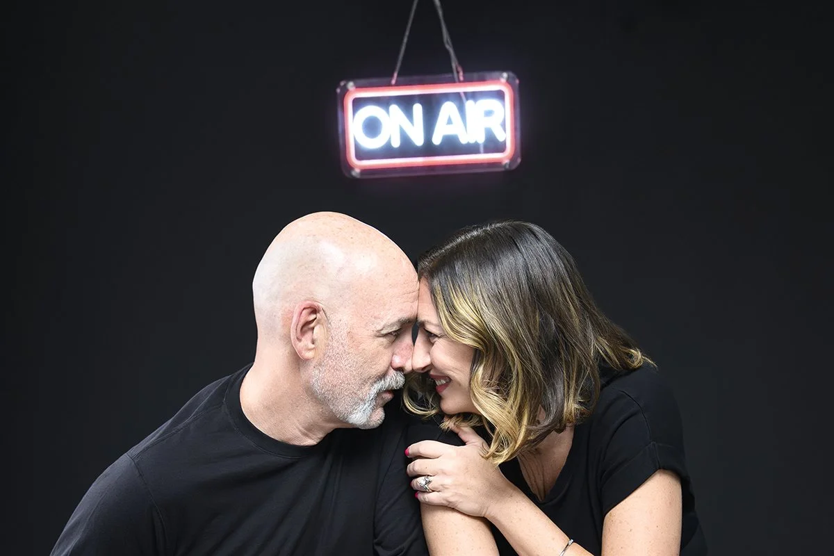 A happy couple with their foreheads touching, smiling, against a black background with an "On Air" sign hanging above them.