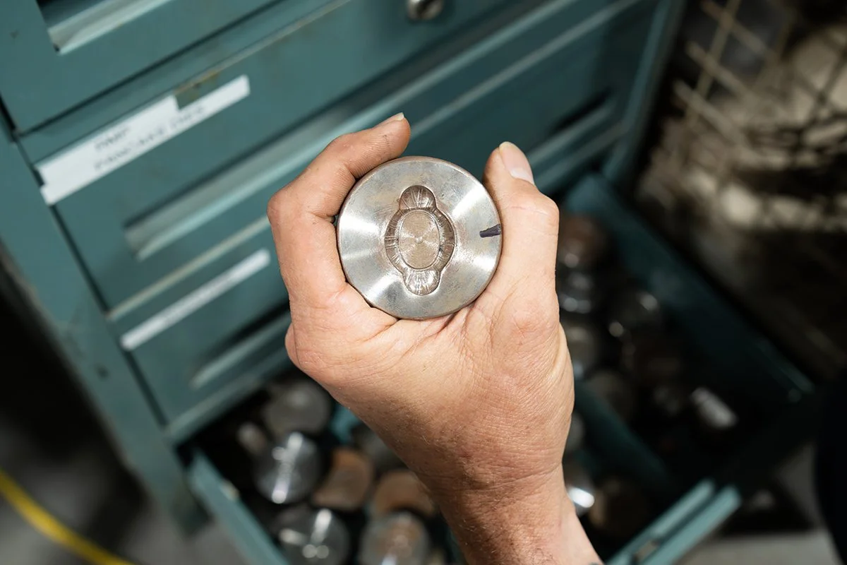 Close-up of a hand holding a metal cylindrical object with a threaded hole in the center, against a background of blue tool drawers and various metal parts.
