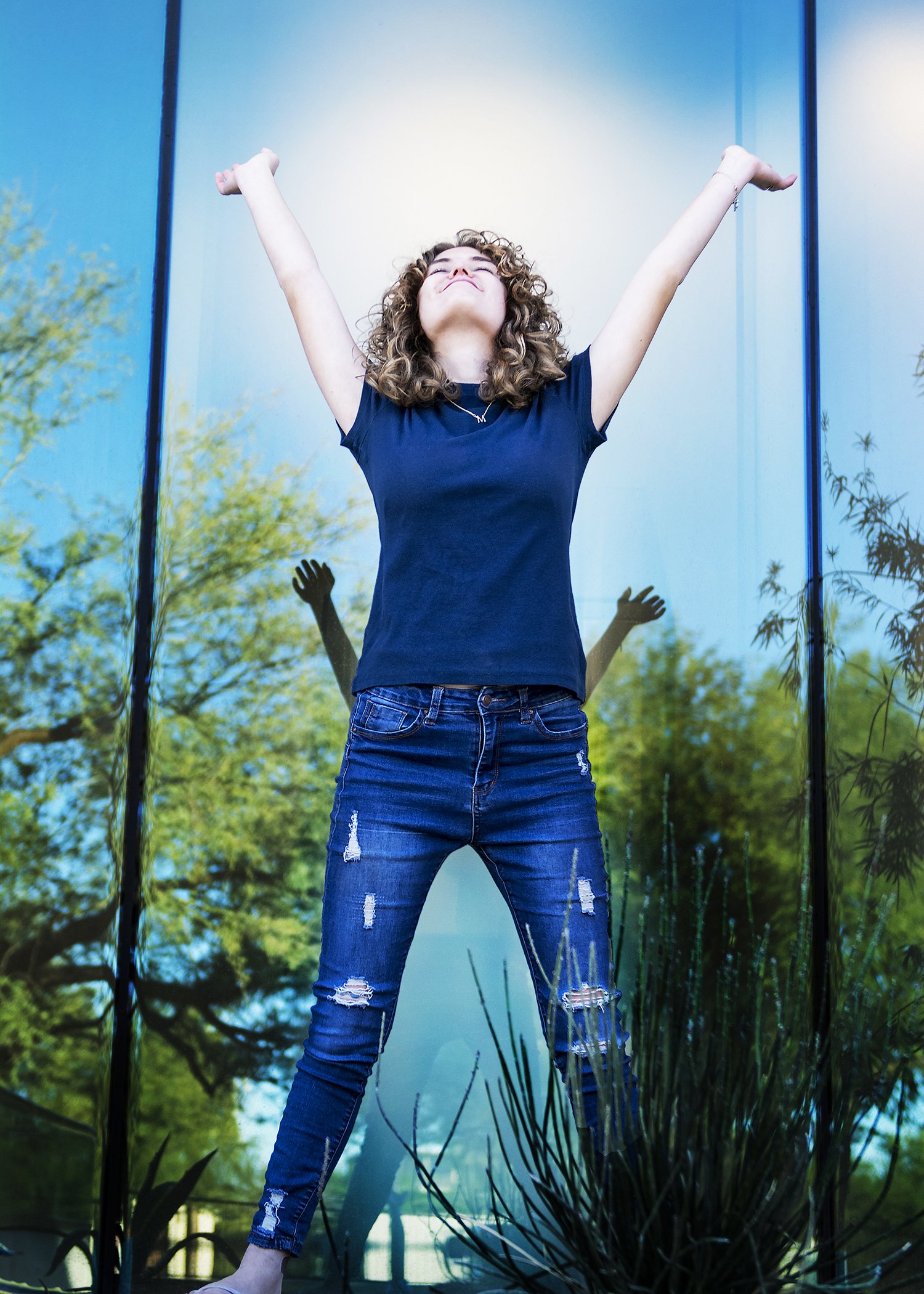 A woman with curly hair, wearing a black t-shirt and ripped jeans, standing outdoors with arms raised in a victory pose in front of a colorful background of trees and water.