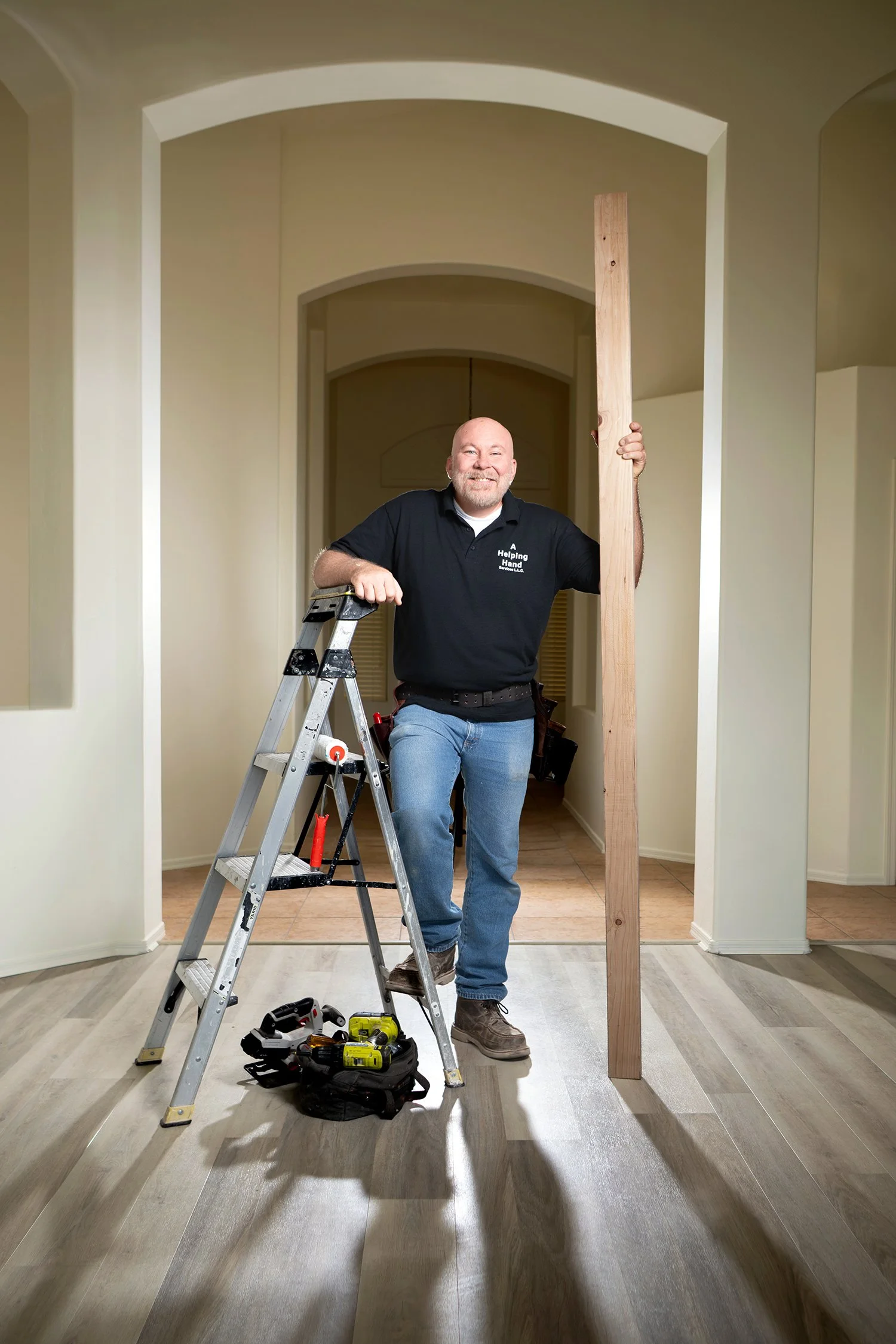 Man standing in a hallway holding a wooden board, with a step ladder, tools, and power drills nearby, preparing to do carpentry work.
