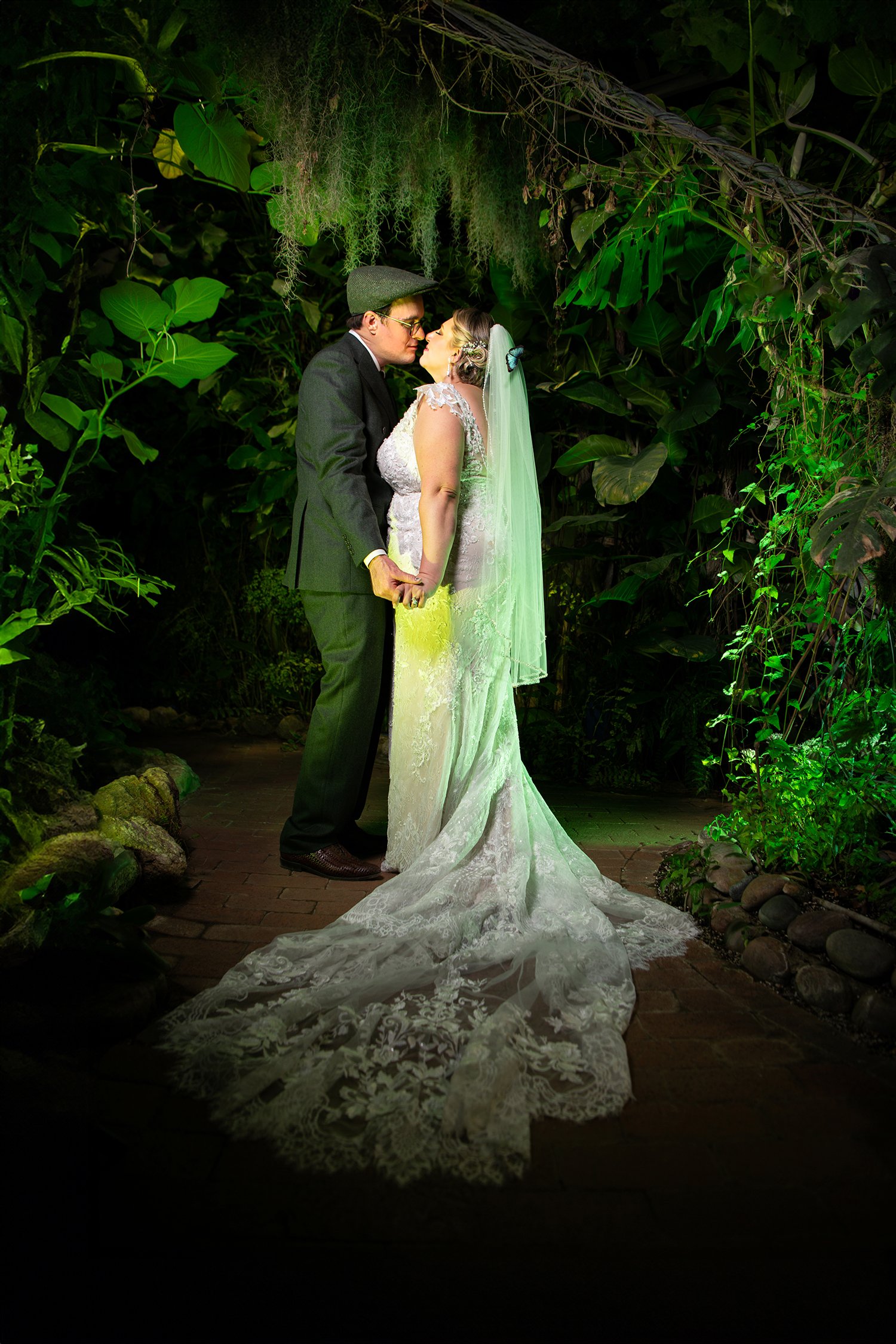 A bride and groom share a kiss in a lush, green tropical garden with large leaves and hanging moss, with the bride in a white lace gown and veil and the groom in a dark suit and glasses.