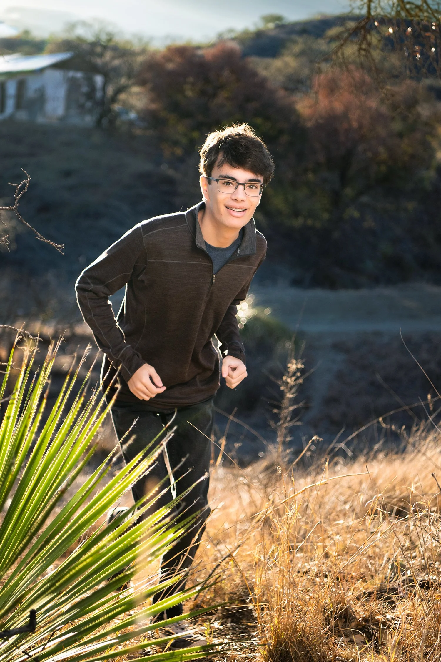 A young man with glasses and dark hair, wearing a dark jacket and dark pants, is outdoors in nature, smiling and running through dry grass with a background of trees and a hillside, illuminated by warm, golden sunlight.