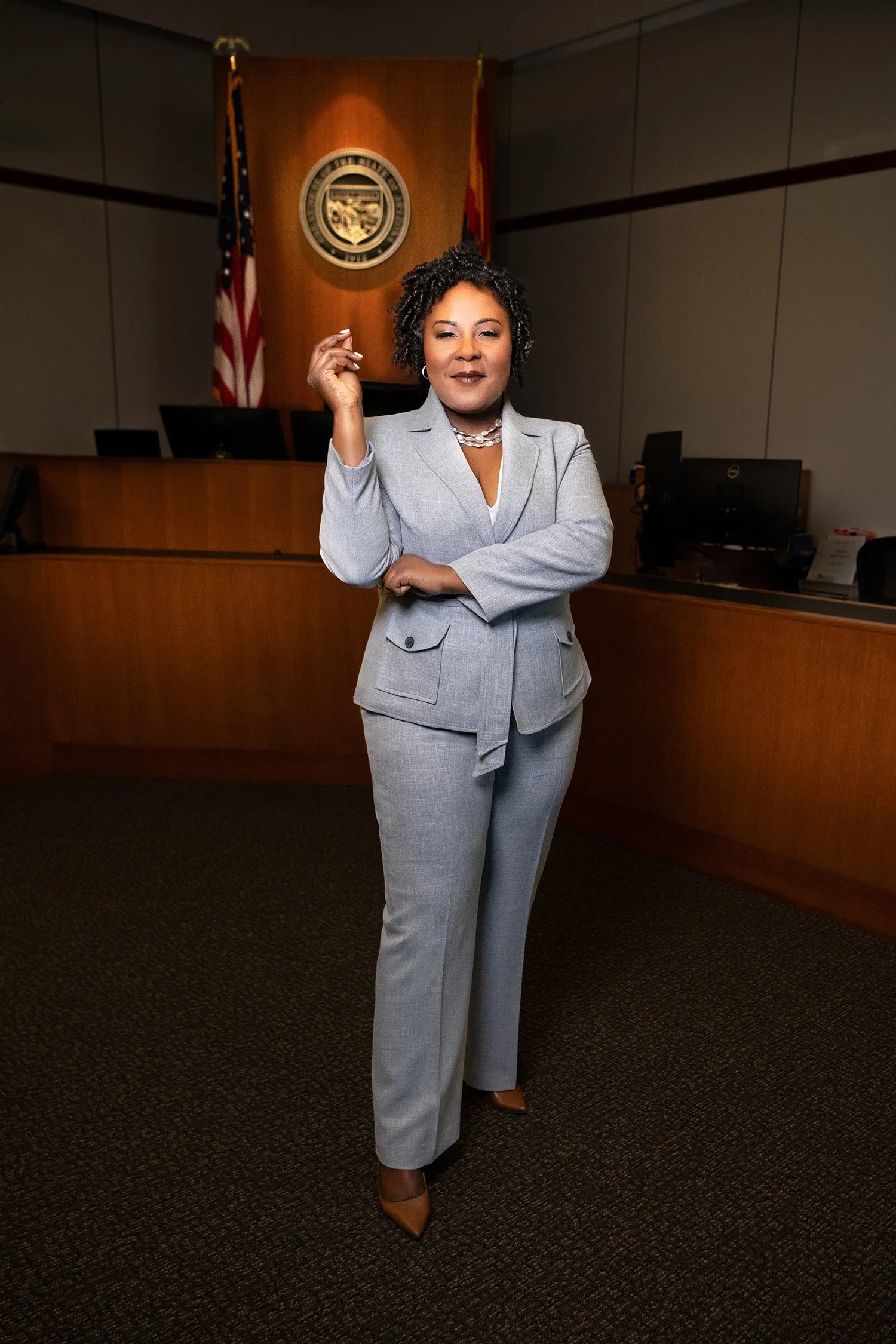 A woman in a gray suit standing in a courtroom, with a wooden judge's bench, a seal on the wall, and two American flags behind her.