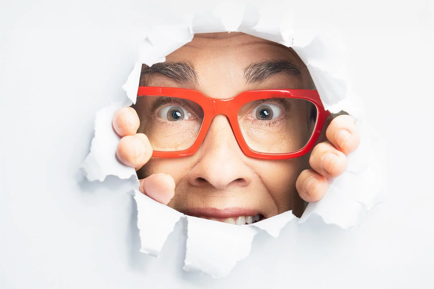 Person peering through torn paper with wide eyes, wearing red glasses posing for a branding photo shoot.