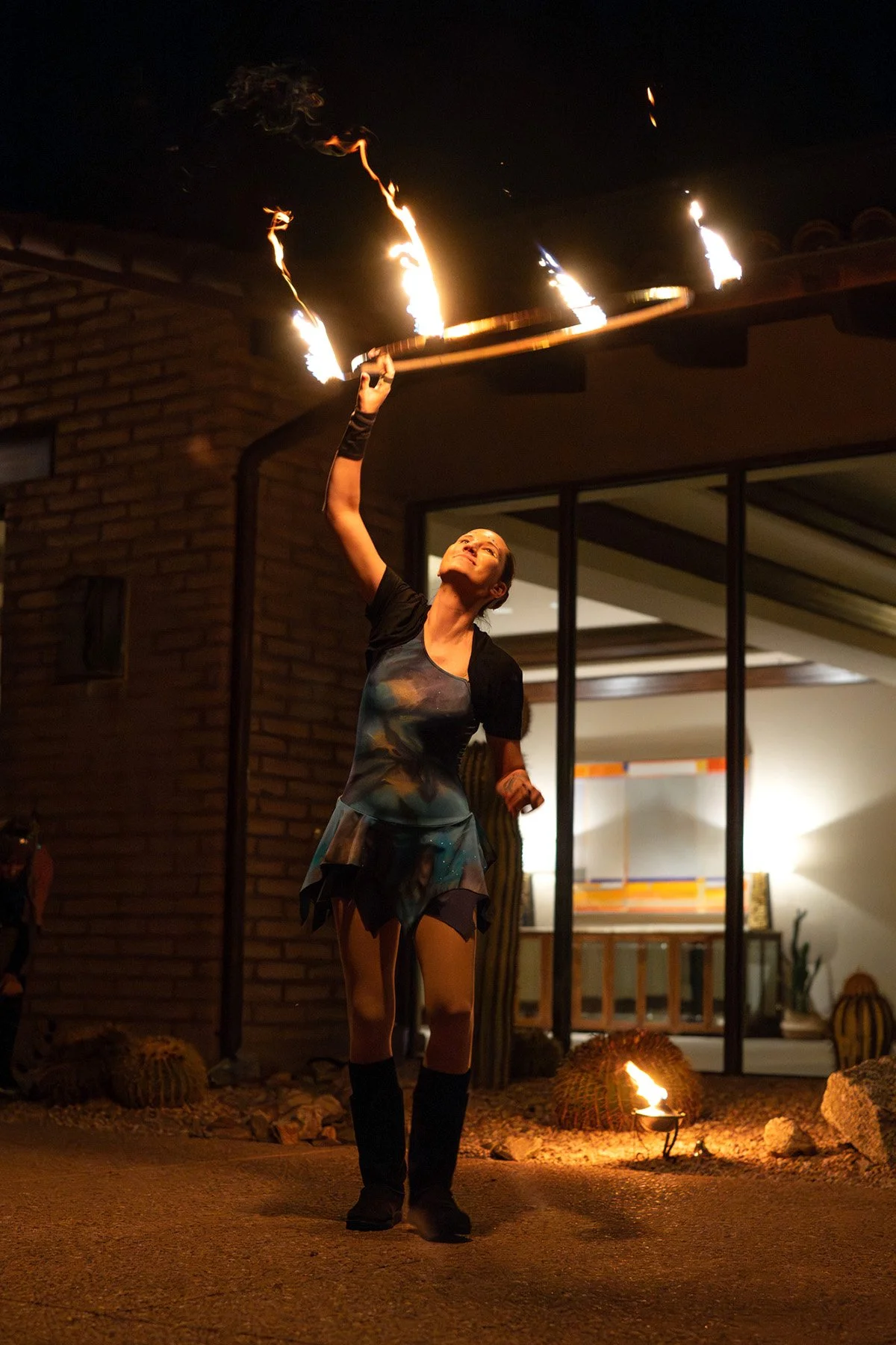 A woman performing fire dancing in front of a modern house at night, with flames on a staff forming sparks in the air, and a small fire on the ground nearby.
