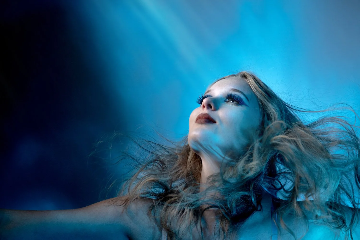 Close-up of a woman with wavy blonde hair, looking upward with a serene expression, illuminated by blue lighting.