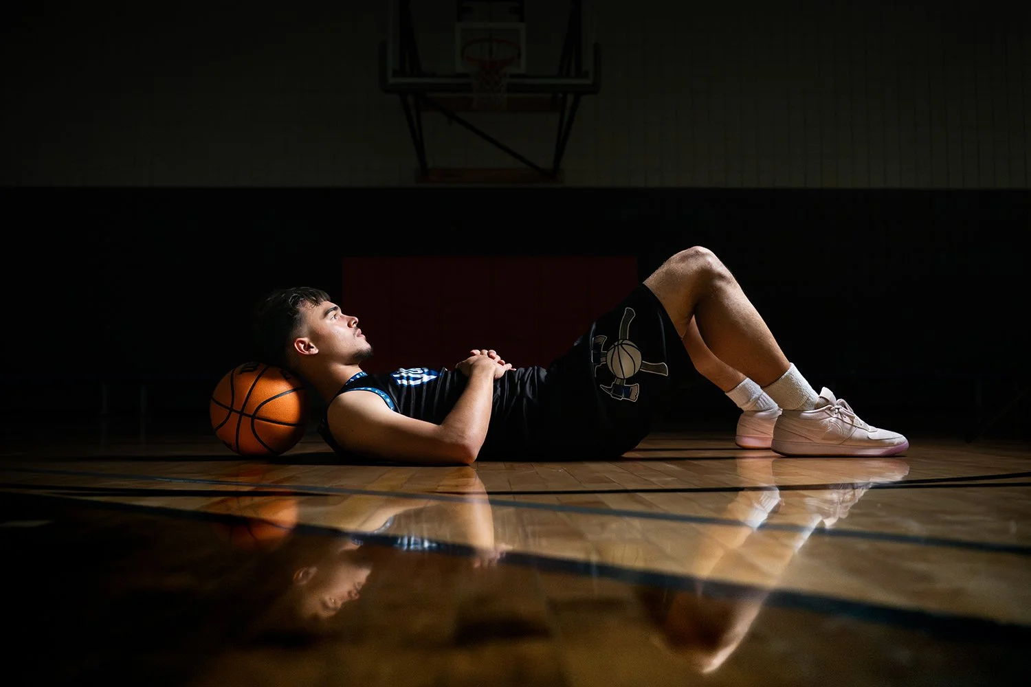 A young male basketball player lying on the hardwood court floor with a basketball under his neck, looking upward in a dark gymnasium.