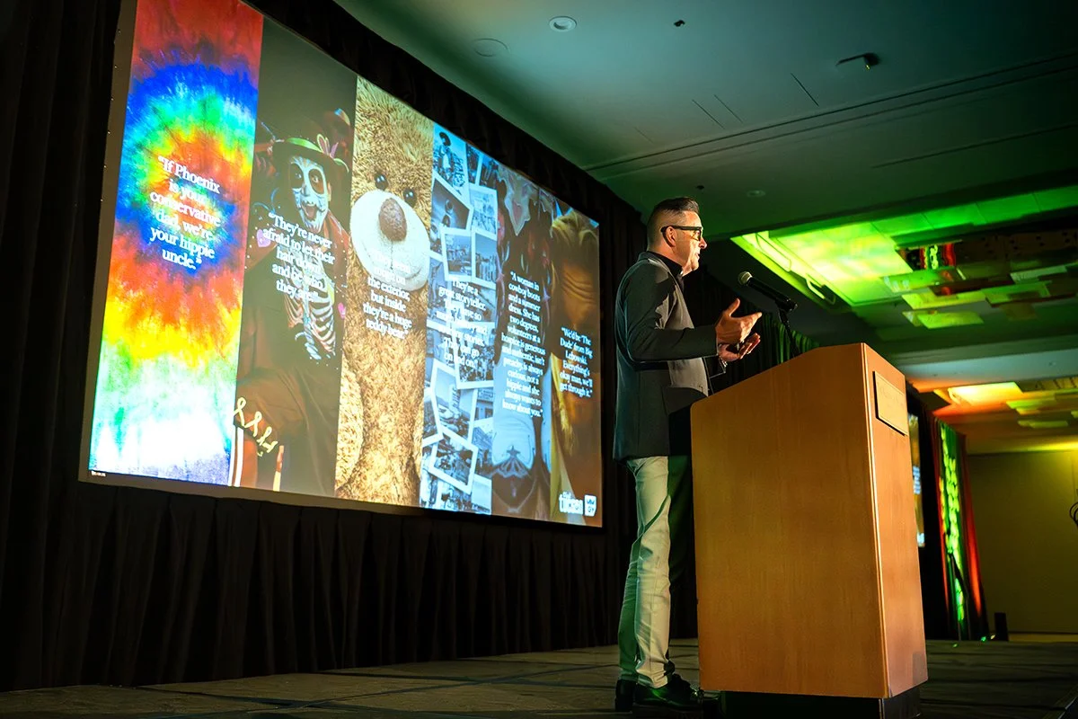 A man giving a presentation at a conference or event. He stands at a wooden podium with a microphone, gesturing with his hands. Behind him is a large screen displaying a colorful collage of images and text, including a tie-dye background with rainbow