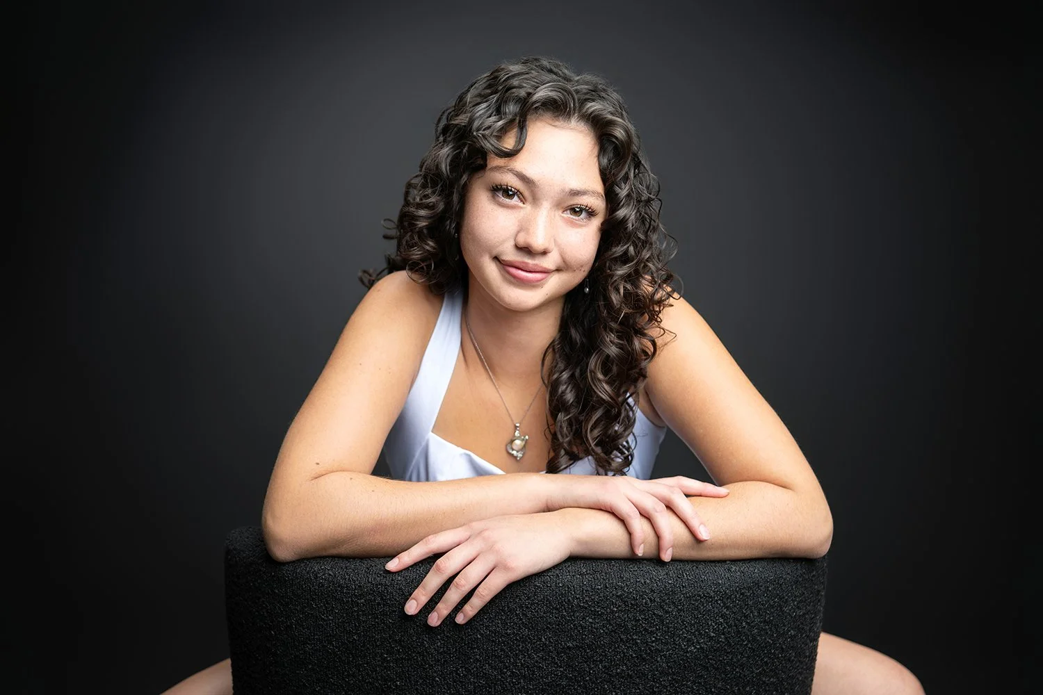 A young woman with long, curly dark hair, smiling, leaning on a black textured surface with her arms crossed, wearing a white sleeveless top and a heart-shaped pendant necklace, against a dark gray background.