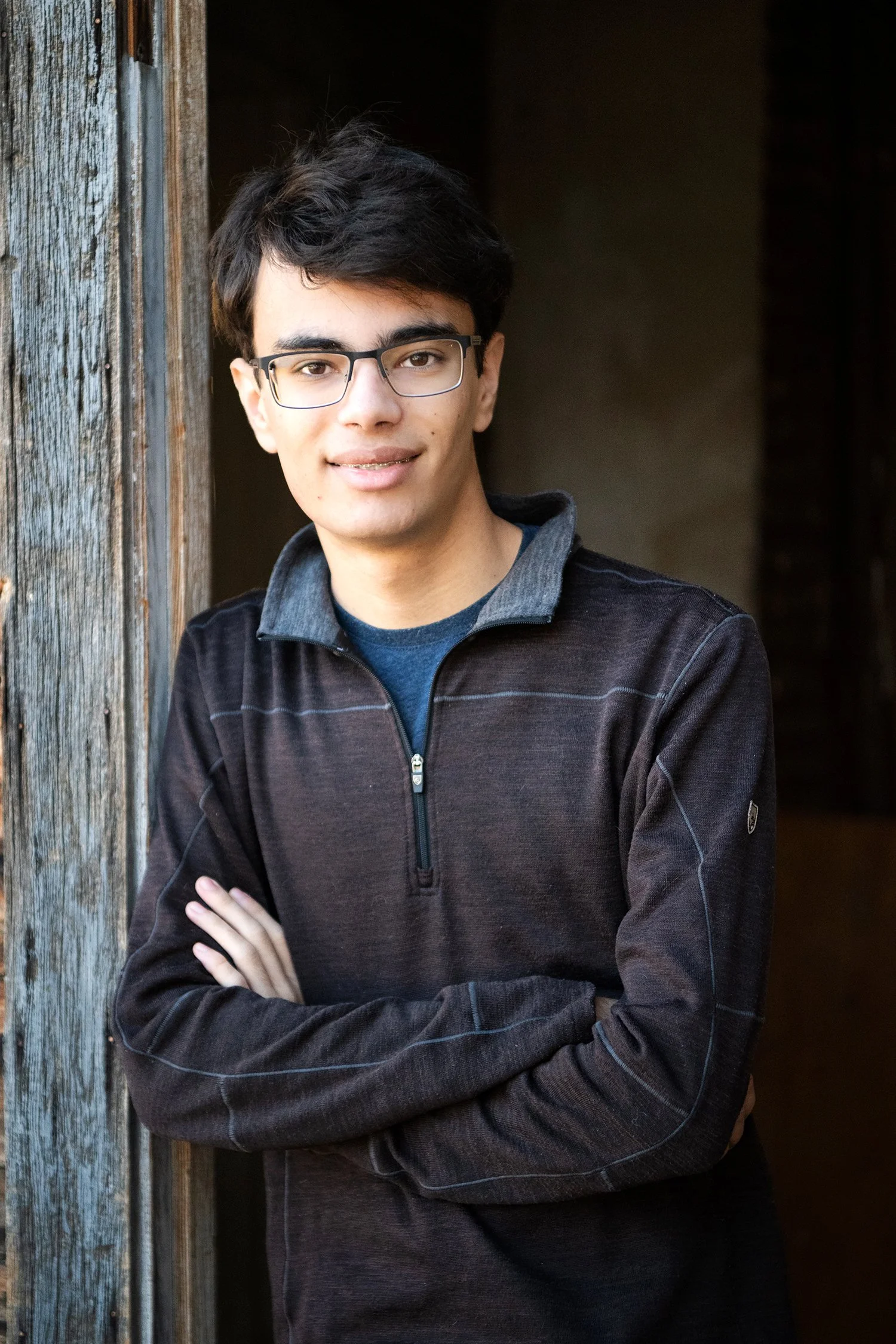 Portrait of a young man with glasses, short dark hair, wearing a dark jacket, standing next to a weathered wooden post with crossed arms.