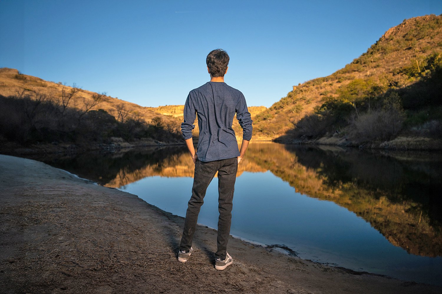 A person stands on the edge of a river with their back to the camera, wearing a blue shirt and black pants, looking at the reflection of surrounding hills in the calm water during sunset.
