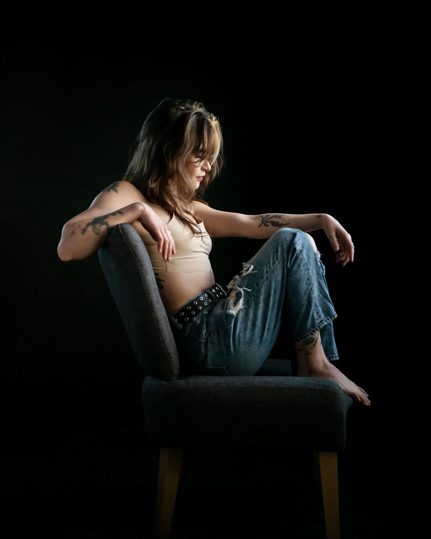 A woman with tattoos on her arms and hair with light and dark brown shades is sitting on a dark-colored upholstered chair with wooden legs. She is wearing a beige crop top and ripped blue jeans. She is barefoot and appears to be lost in thought, with