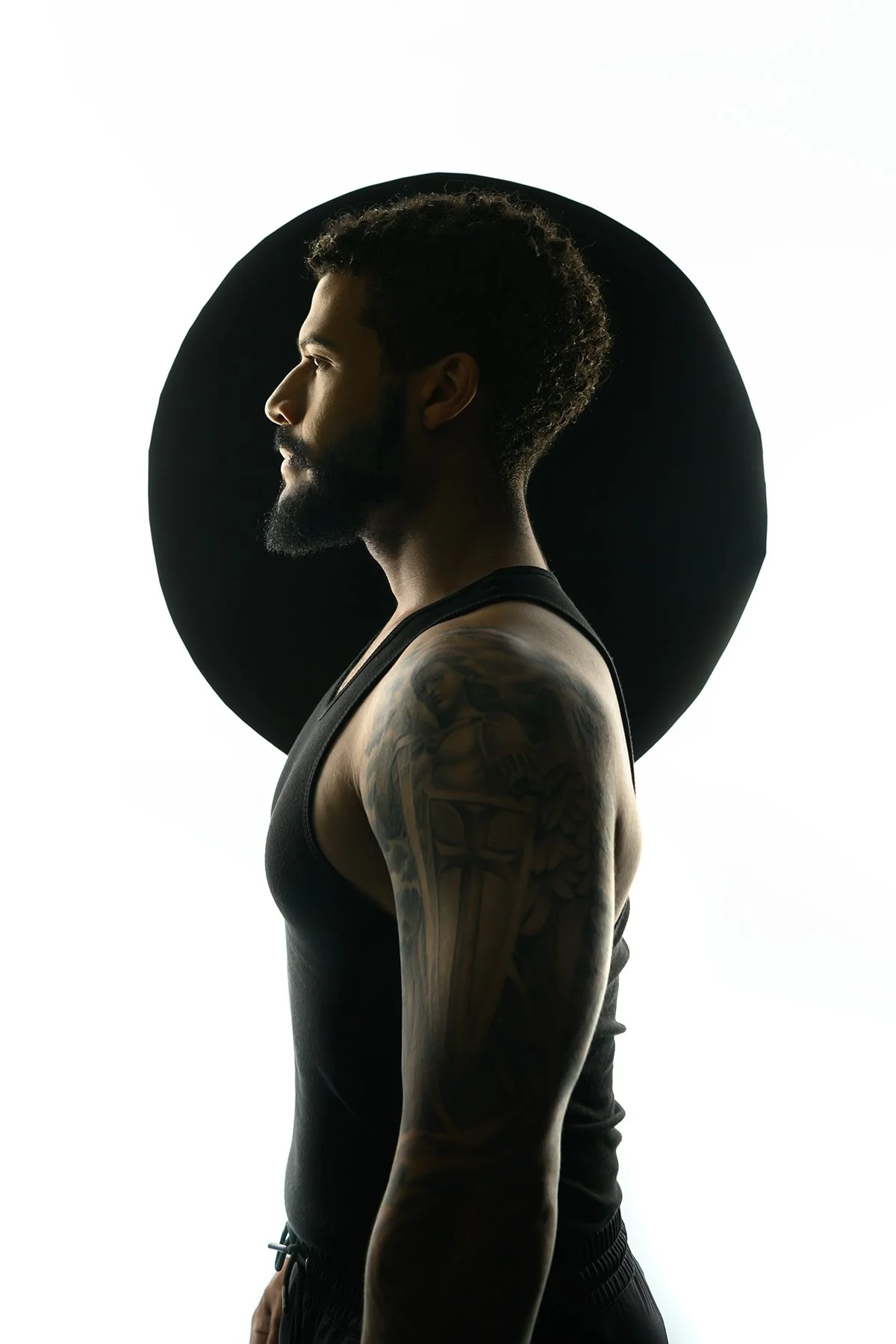 Profile of a tattooed man in a black tank top standing in front of a white background with a black circle.
