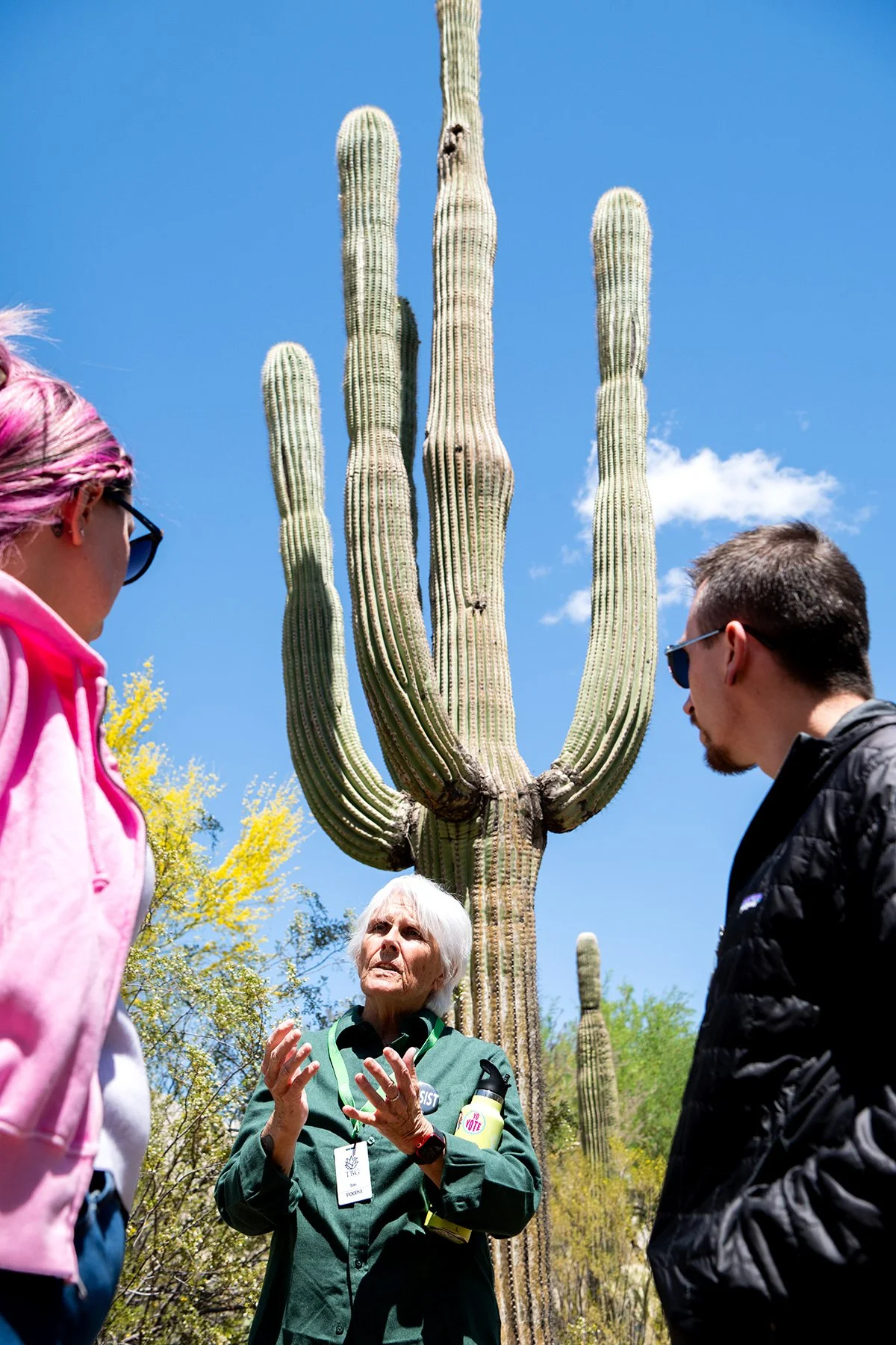 A group of people listening to a woman speak outdoors in front of a tall saguaro cactus, with a bright blue sky in the background.