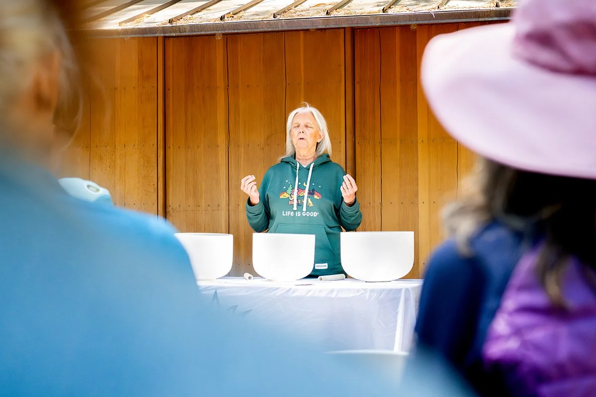 A woman with gray hair in a teal hoodie, standing behind a table with white bowls, appears to be speaking or performing in front of an audience in a wooden-paneled room, with two people in hats and jackets in the foreground.