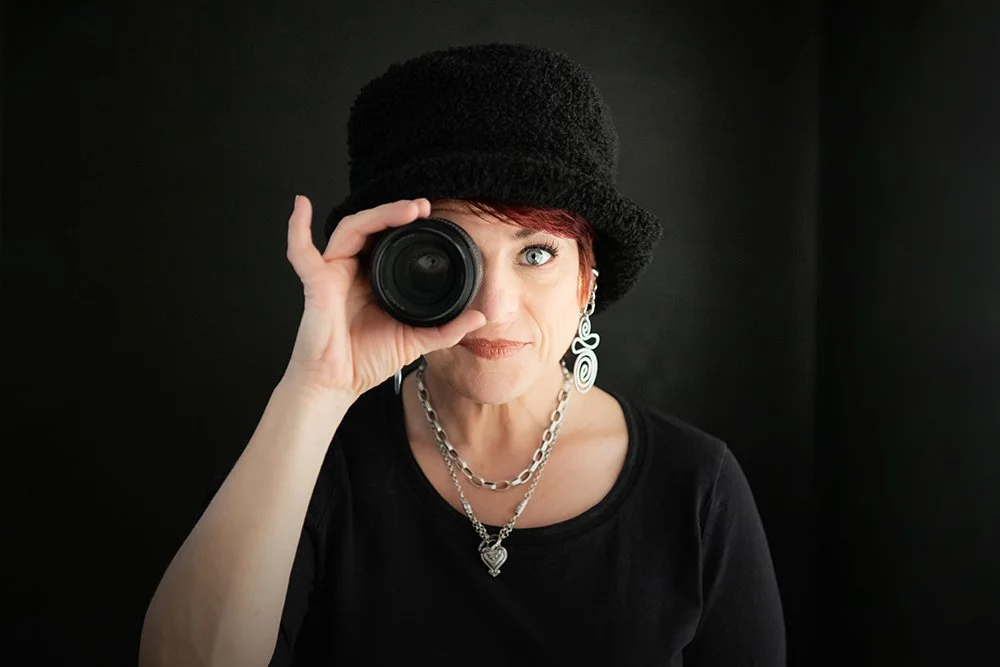 Photographer Amy Ijams with red hair, wearing a black hat and earrings, holding a camera lens up to her right eye, against a dark background.