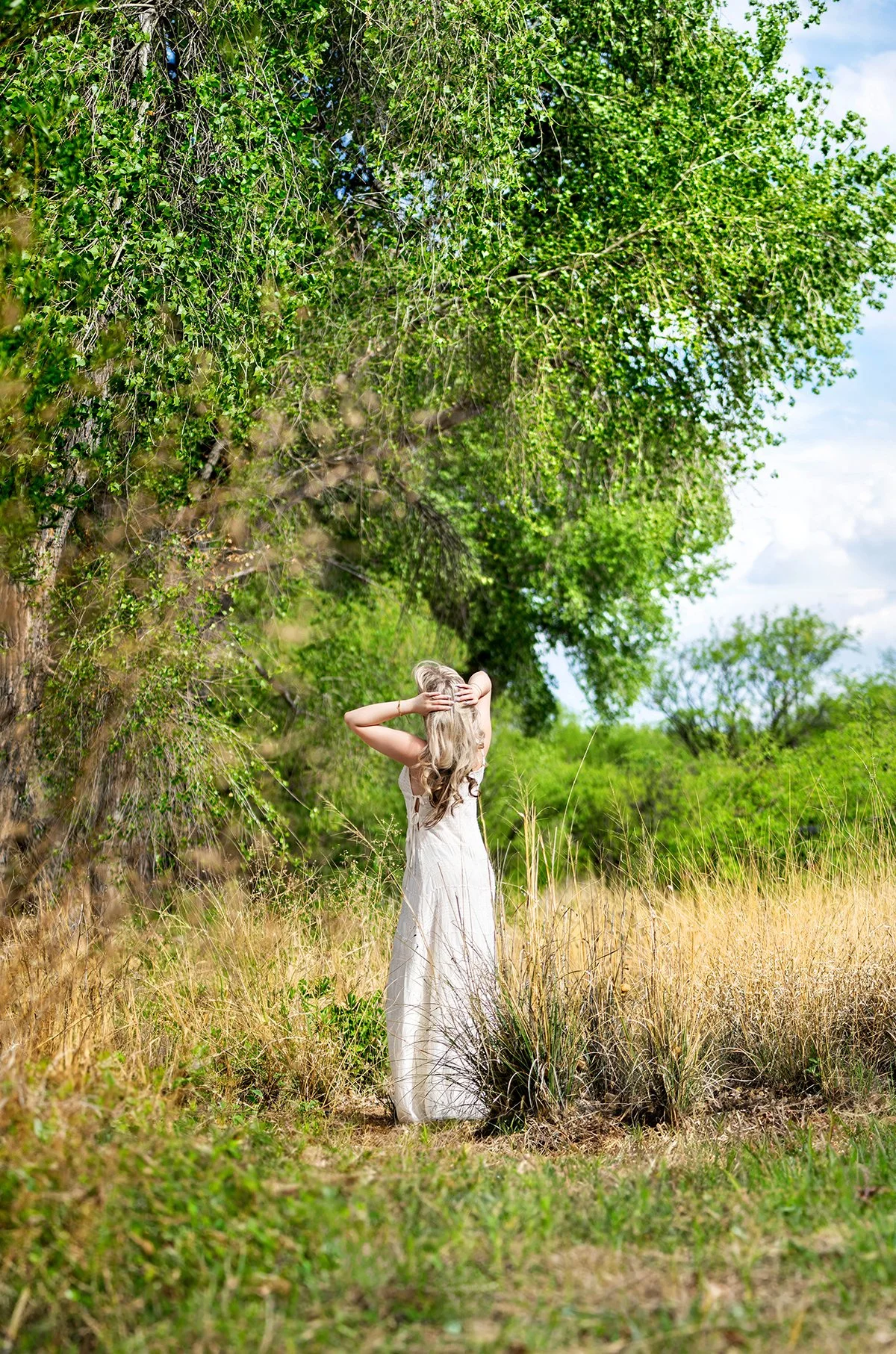 A Tucson high school senior in a white dress standing in a grassy field with tall grass and bushes, reaching up to touch her head, with large trees and a blue sky in the background during a photo shoot