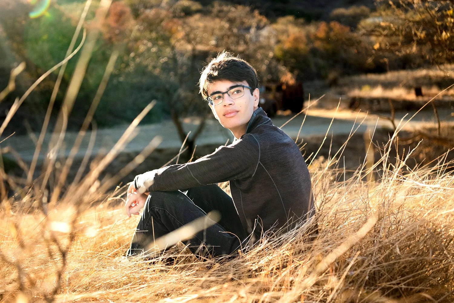 A young man with glasses sitting in a field of tall, dry grass during sunset, with trees and a hill in the background.
