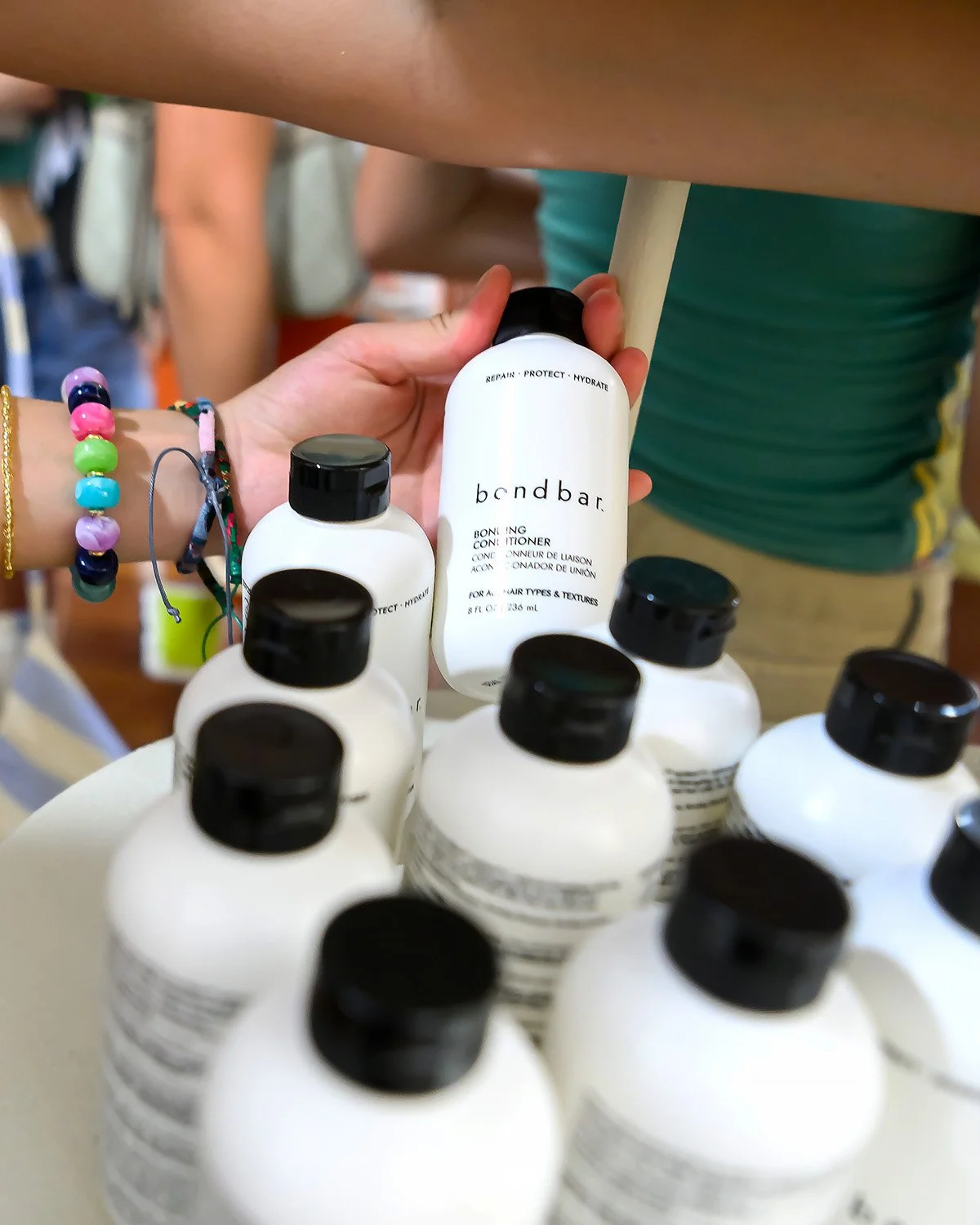 Multiple white bottles of hair conditioner on a display, with one bottle being held by a person wearing colorful beaded bracelets. The bottles are labeled "bendbar" with additional text.