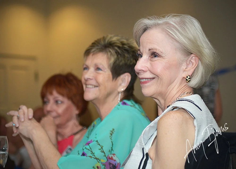 Three women sitting at a table, smiling and clapping during an event. The woman in the foreground has short gray hair and is wearing a sleeveless top with earrings and a necklace. The woman in the middle has short, layered hair and is wearing a teal 