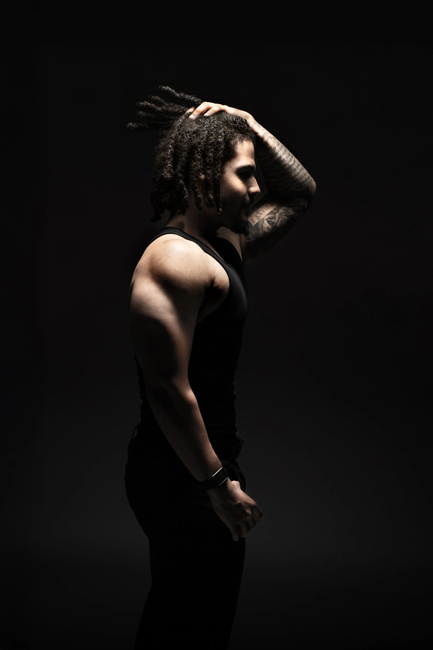 Side profile of a man with dreadlocks, tattoos on his arms, wearing a black sleeveless shirt, against a dark background.