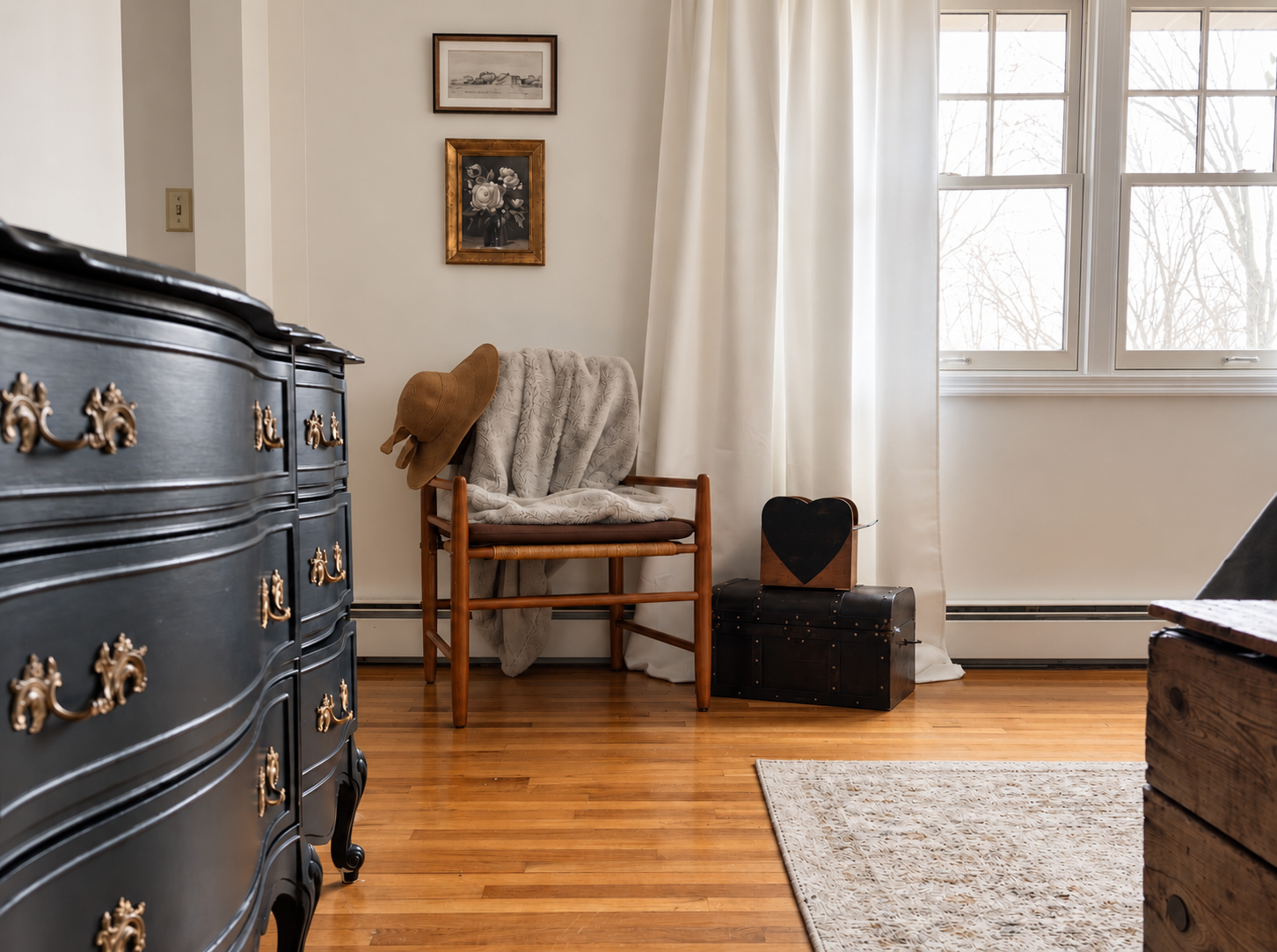 Corner of a room with a black antique dresser with brass pulls, a wood chair and hardwood floors