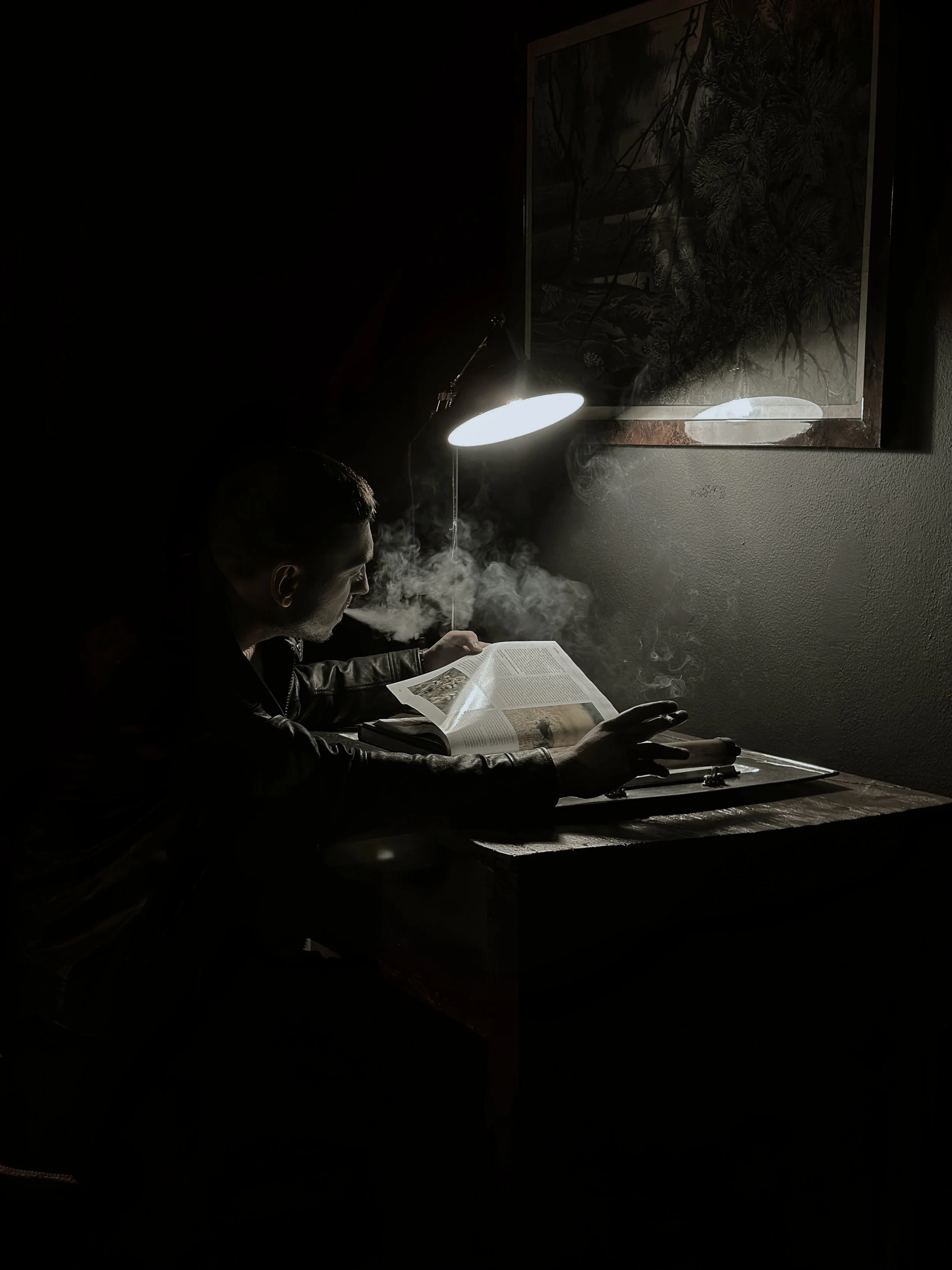 A person sitting at a wooden table in a dark room, reading a book under a bright desk lamp. Smoke rises from an unseen cigarette, illuminated by the lamp.