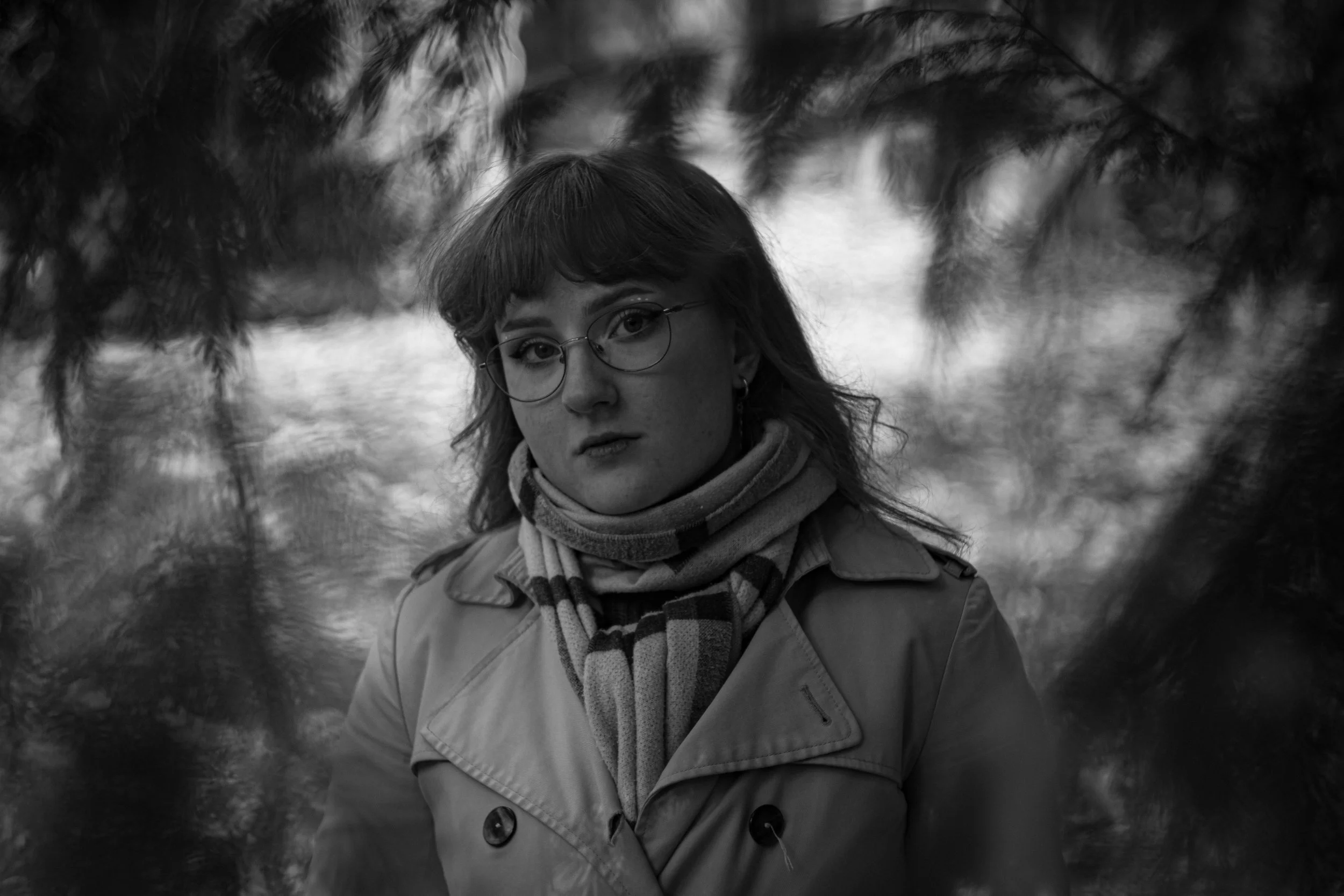 Schwarz-Weiß-Foto eines Mädchens mit Brille und Schal in einer natürlichen Umgebung mit Bäumen.