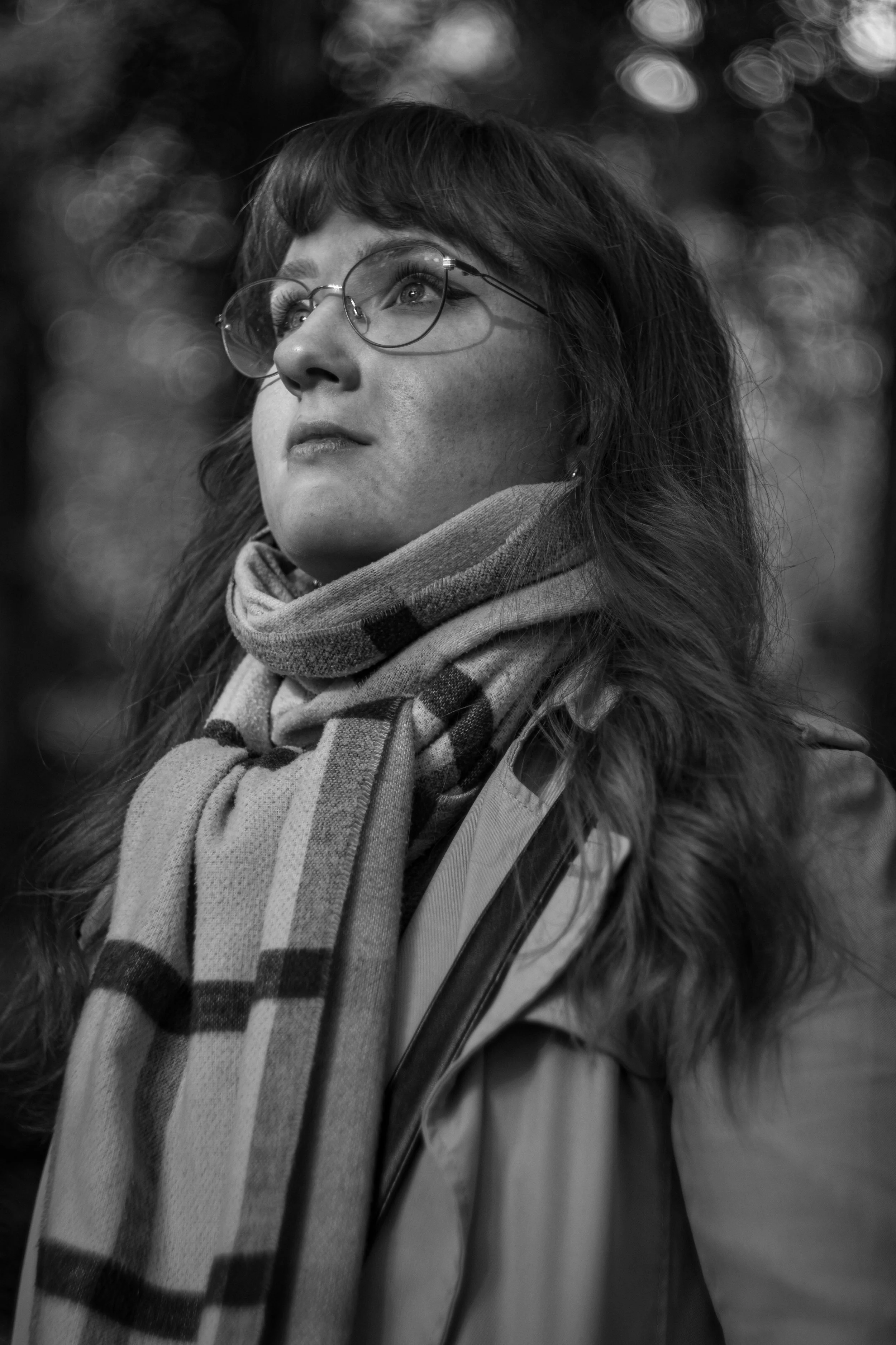 Schwarz-weiß-Fotografie eines jungen Mädchens mit Brille, das nach oben schaut, mit einem karierten Schal und einer Jacke mit Fellkapuze, im Hintergrund unscharfe Lichter.