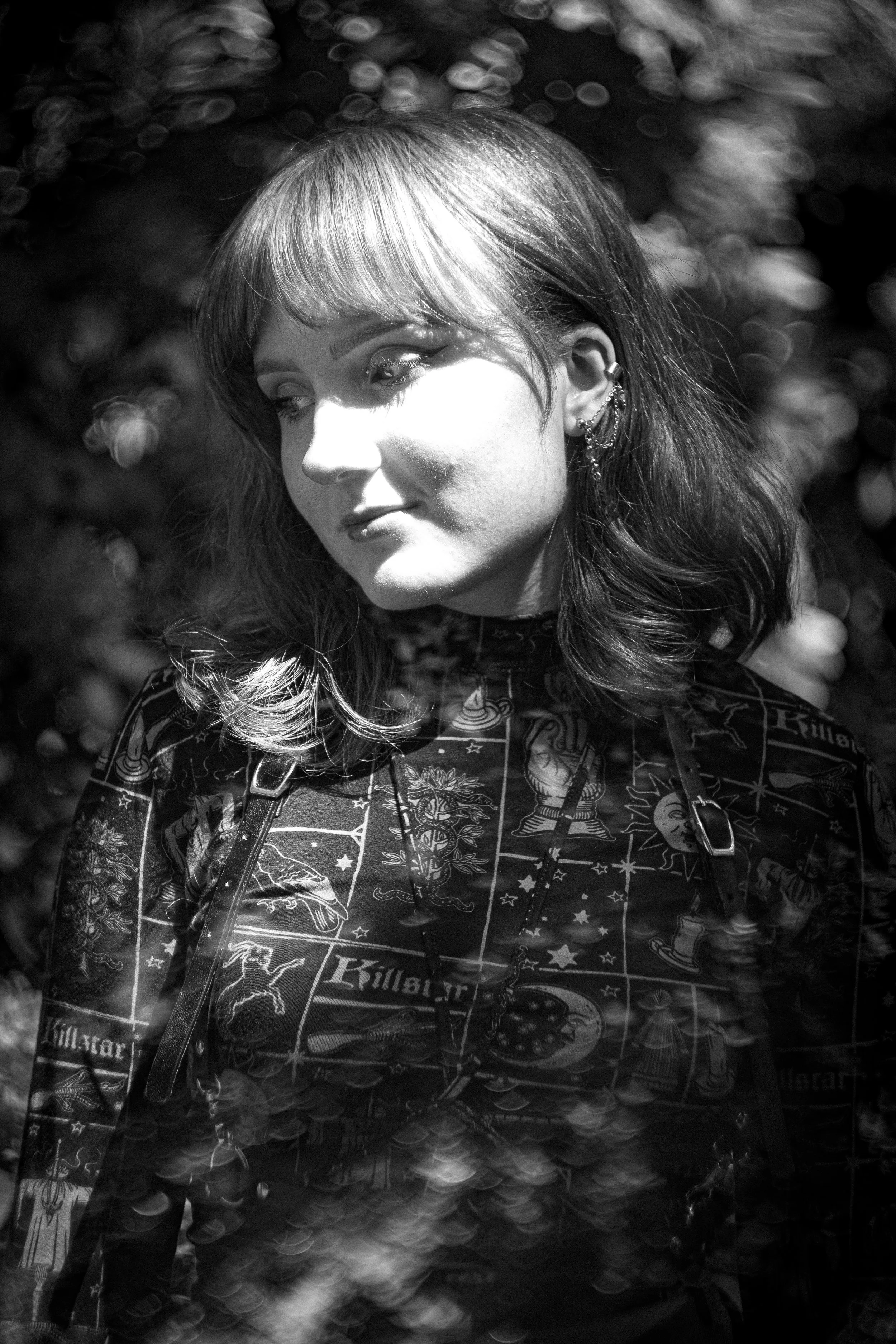 Ein schwarz-weiß Portrait einer jungen Frau mit dunklen Haaren und Piercings am Ohr, die nach rechts schaut, vor einem Hintergrund mit Lichtreflexen.
