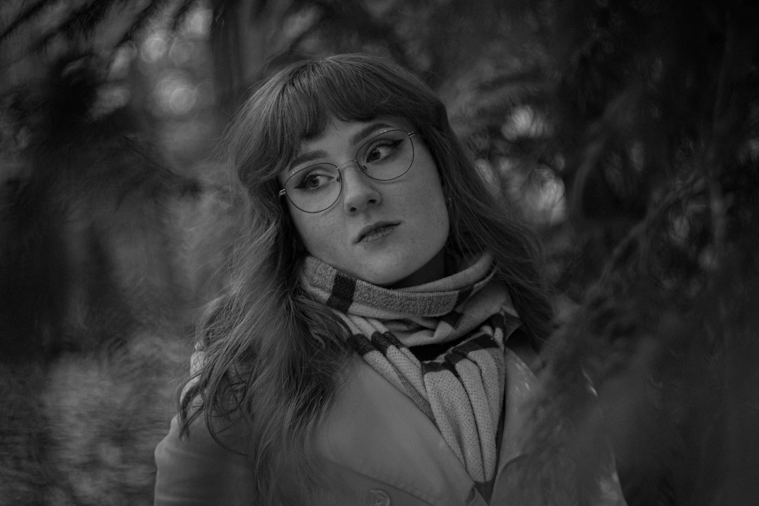 Junge Frau mit Brille, scarf und langen Haaren schaut nach links, im Hintergrund unscharfe Natur.