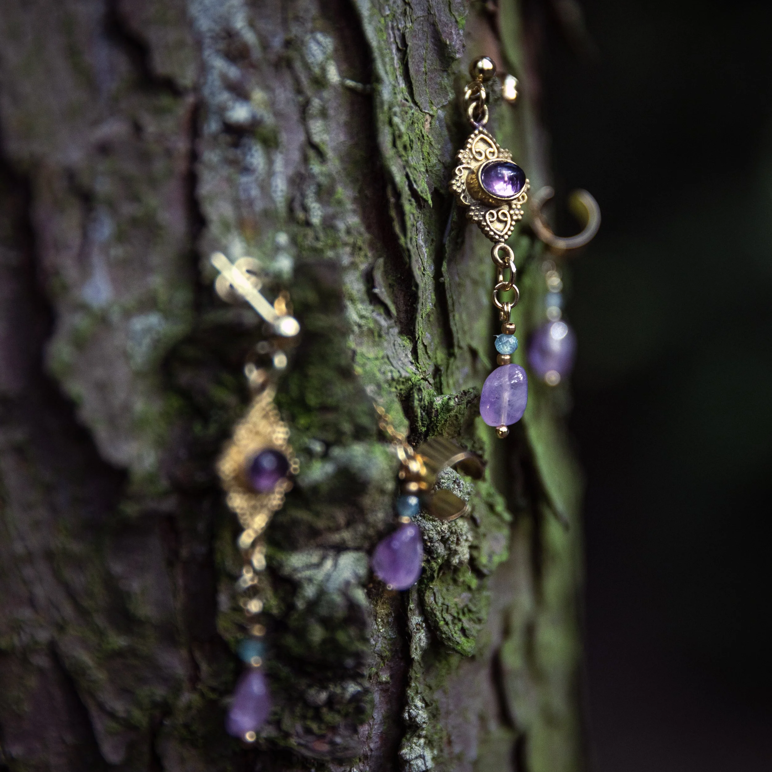 Eine goldene Halskette mit violettem Edelstein hängt an einem Baum mit grober, moosbedeckter Rinde.
