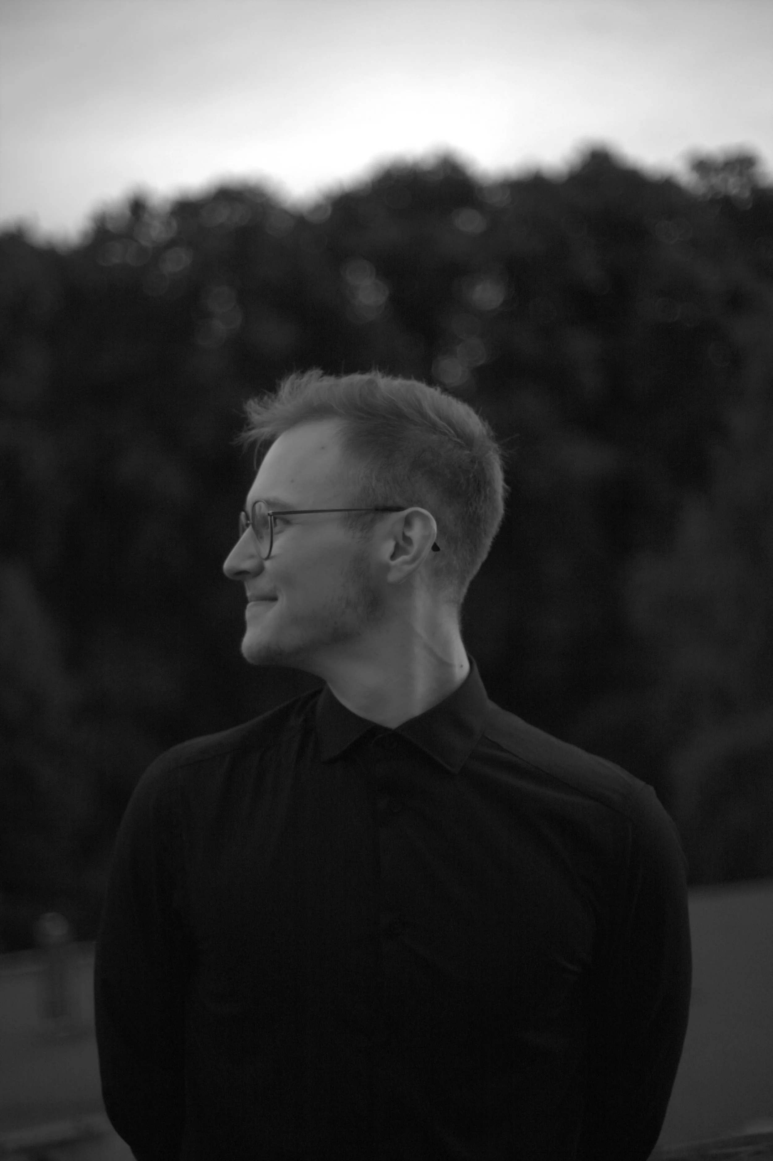 Junge Person mit Kurzhaarschnitt und Brille, trägt schwarzen Pullover, lachend, im Freien bei Sonnenuntergang, unscharfer Hintergrund mit Bäumen.