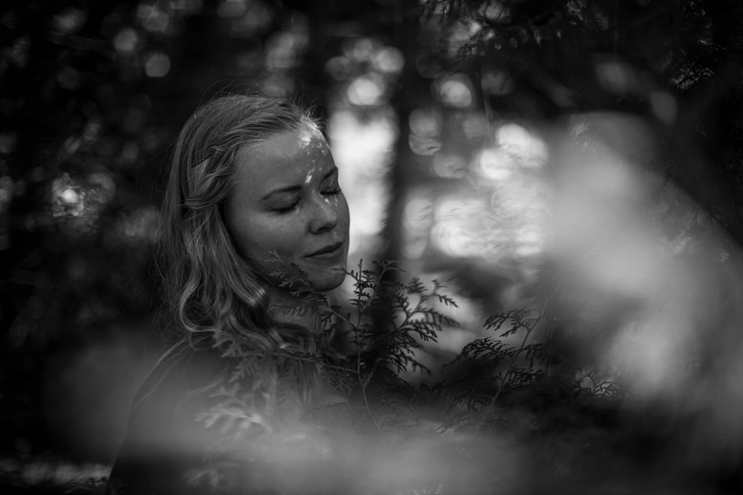 Ein schwarz-weiß Foto eines lächelnden Mädchens mit geschlossenen Augen, umgeben von Blättern und Bäumen in einem Wald.