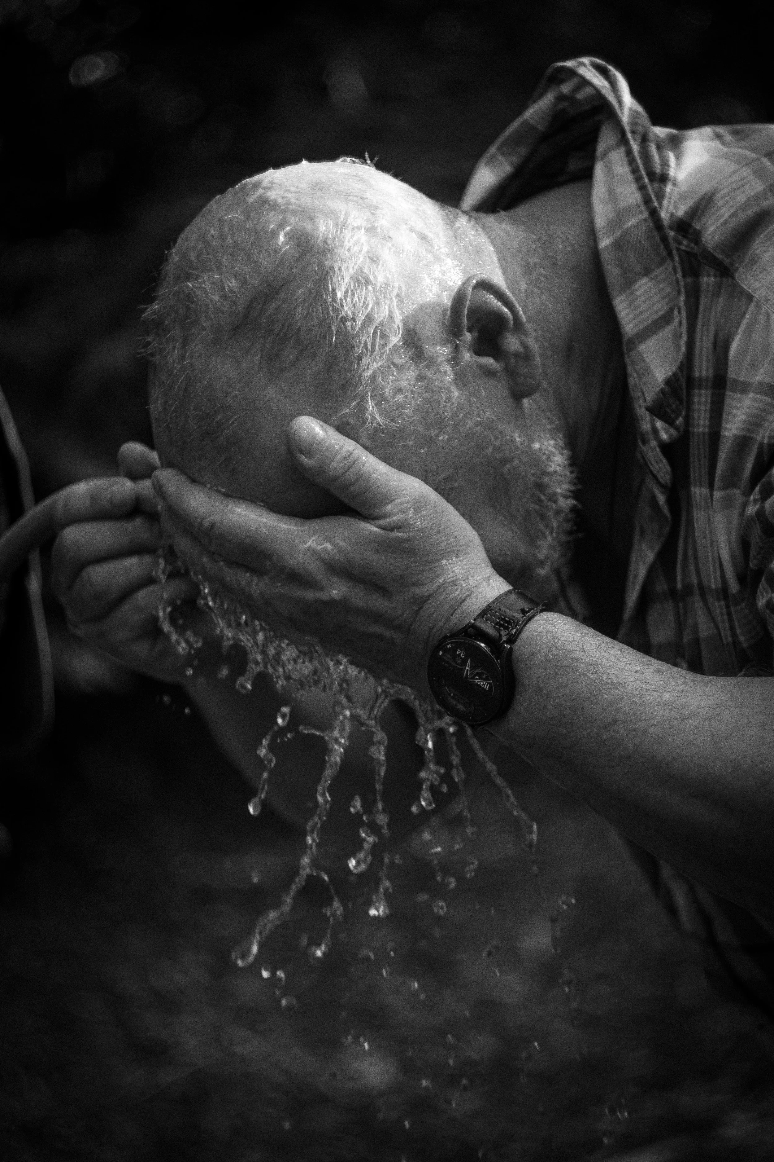 Nahaufnahme eines älteren Mannes mit grauem Bart, der Wasser über den Kopf schüttelt, was Wasser sprudelt, mit einem Armband und einer Uhr am Handgelenk, im Freien.