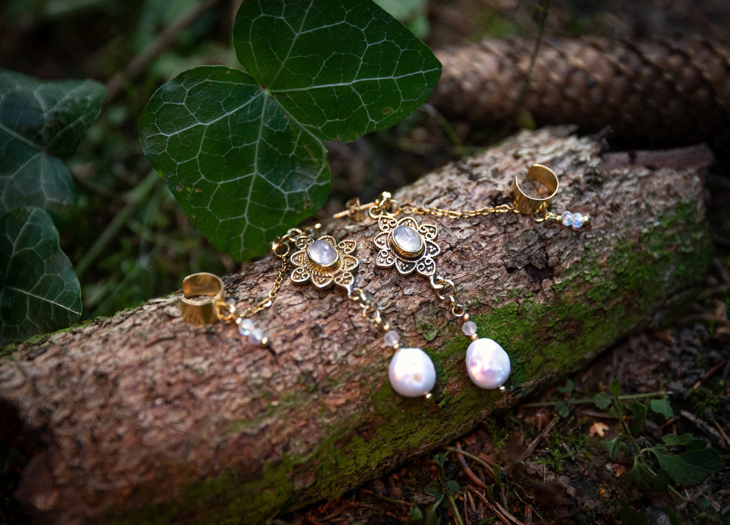Goldschmuck, einschließlich Ohrringe und Kette, liegen auf einem Baumstamm im Wald, umgeben von grünen Blättern.