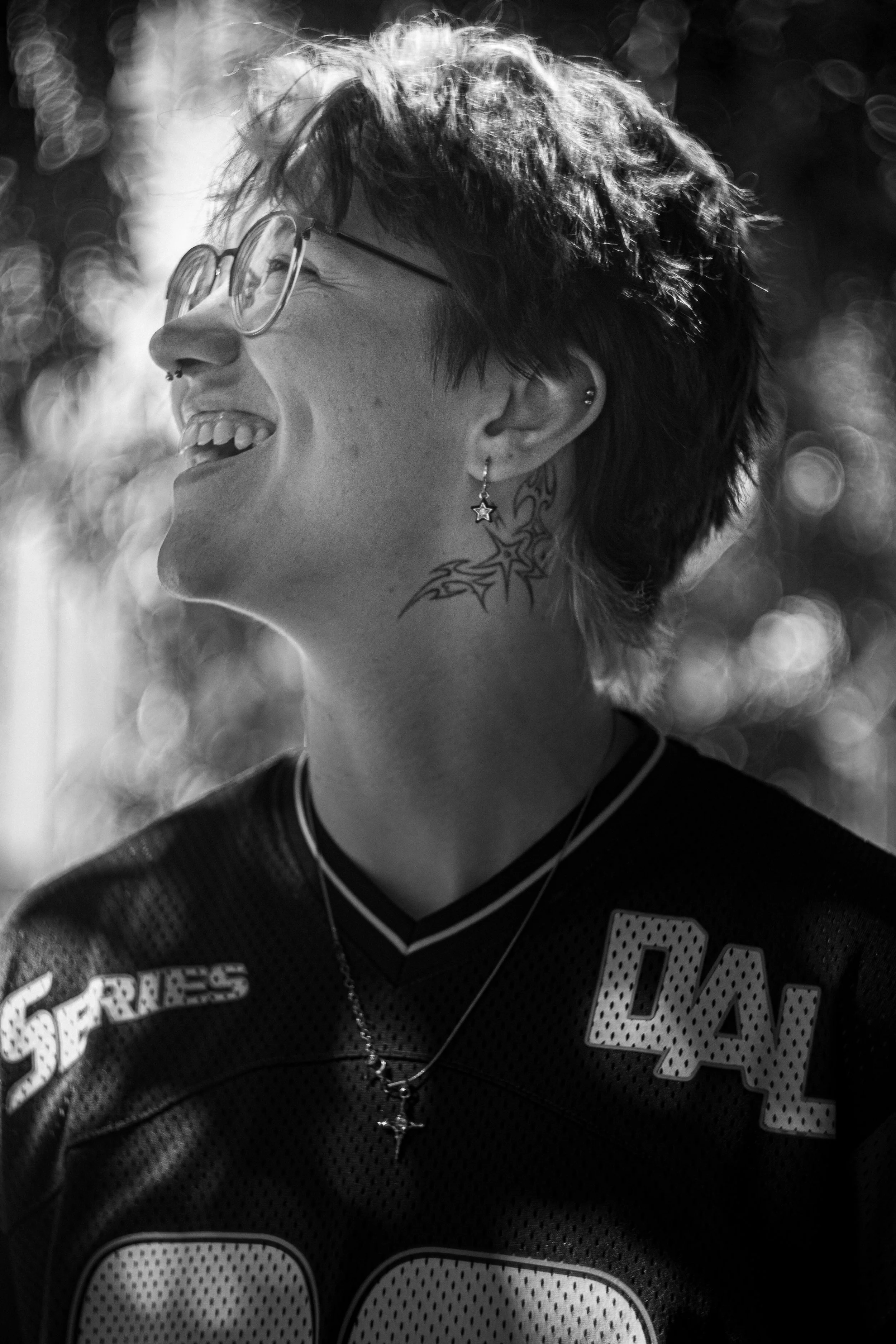 Schwarz-weiß-Foto eines lachenden jungen Mannes mit Kurzhaarfrisur, Brille, mehreren Tattoos, Ohrringen und Halsschmuck. Trägt ein dunkles Trikot mit weißen Nummern und Buchstaben.