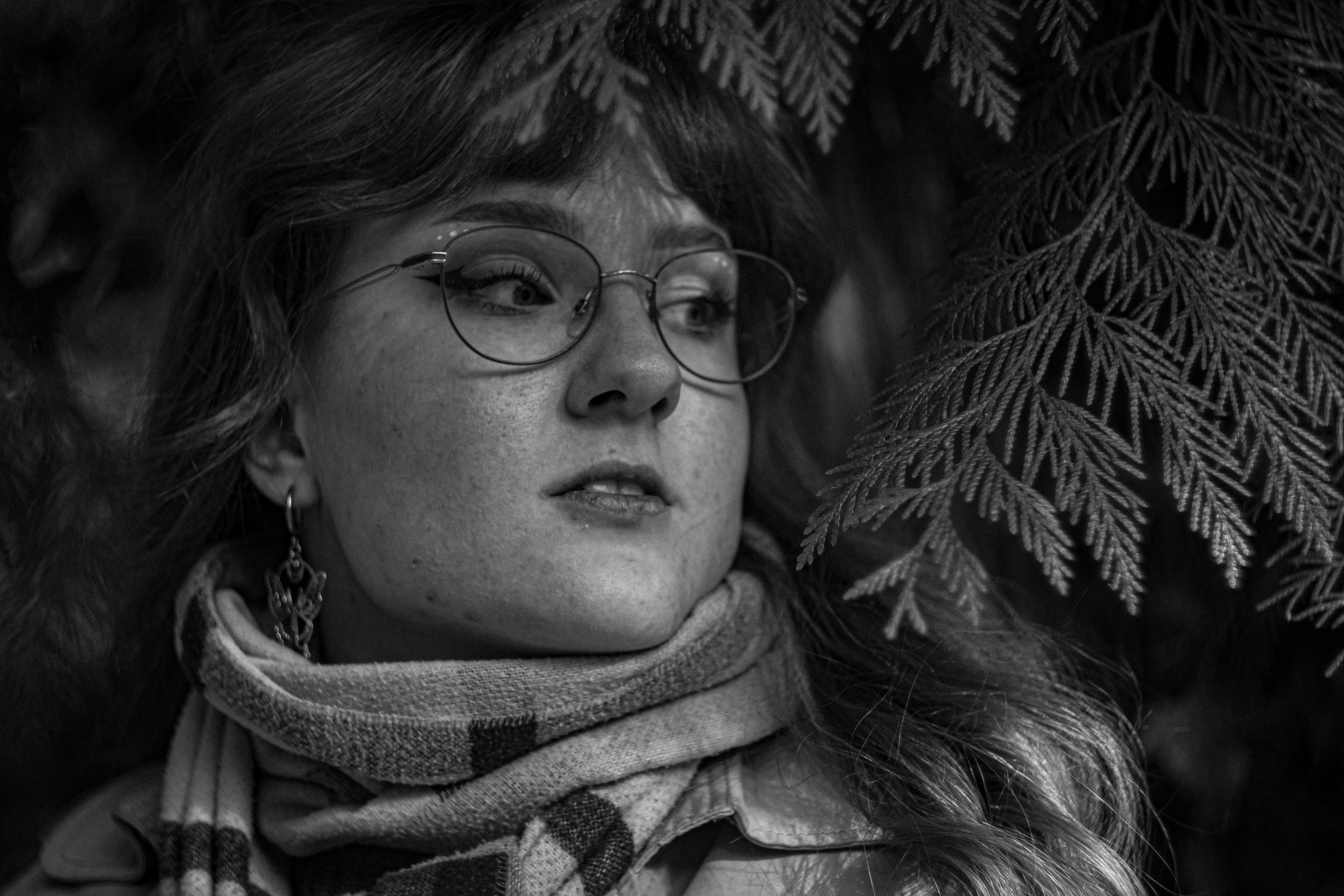 Schwarz-weiß-Fotografie eines jungen Mädchens mit Brille, das zwischen Tannenzweigen liegt.