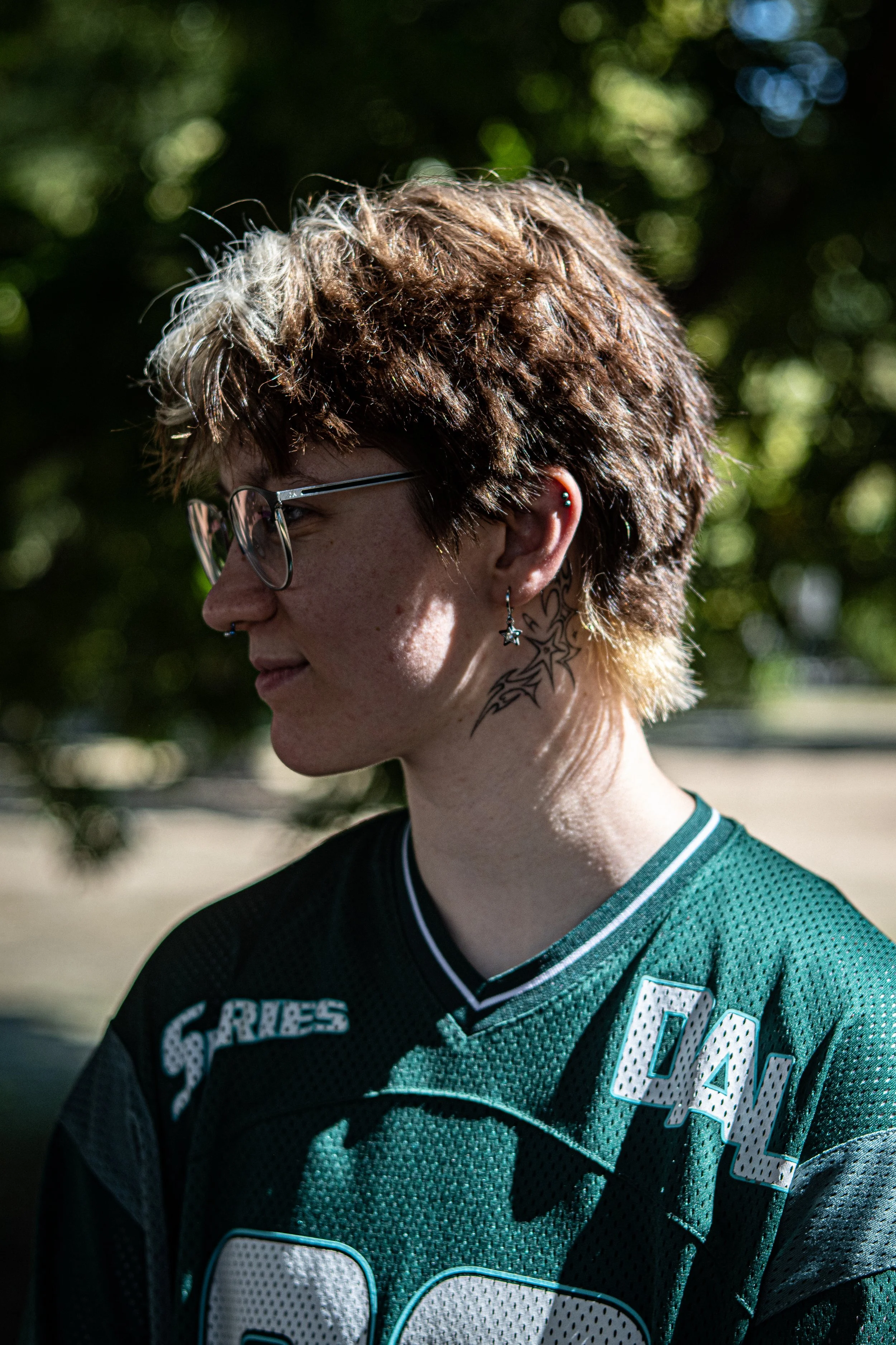Porträt einer jungen Frau mit kurzen, lockigen braunen Haaren und Brille, die in einem grünen Sporttrikot steht, im Hintergrund unscharfe grüne Blätter.
