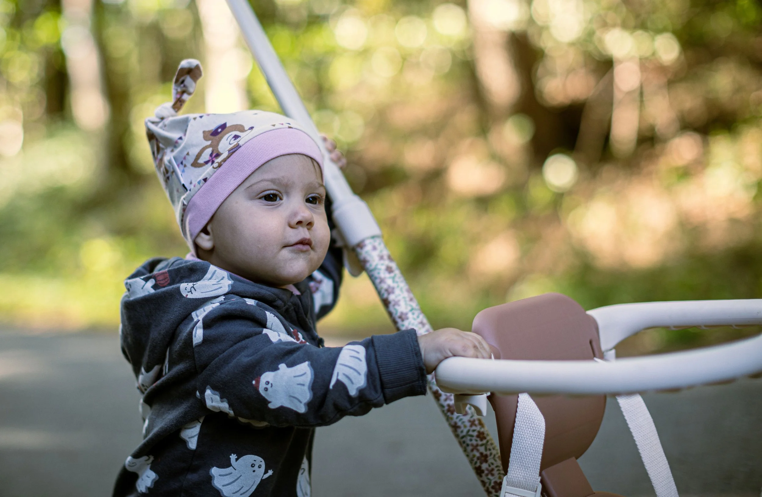 Kleines Kind in einem Kinderwagen in einem Park mit Bäumen im Hintergrund, trägt eine Mütze mit Hase-Design.
