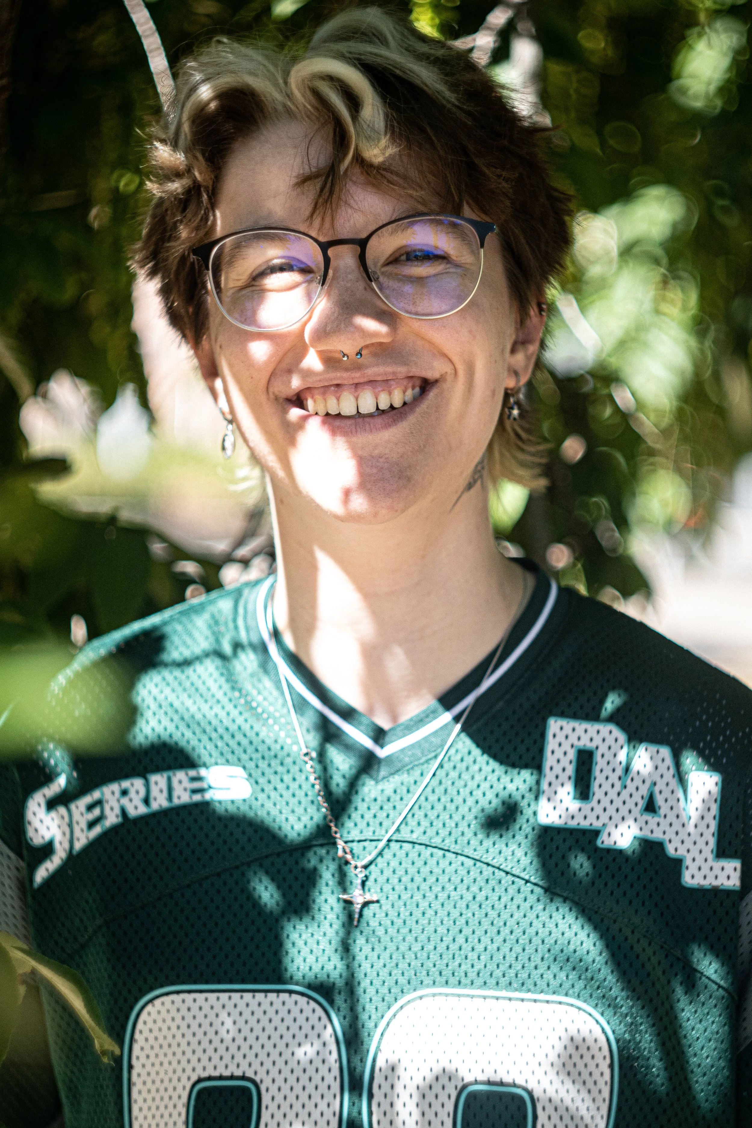 Nahaufnahme eines lachenden jungen Mannes mit Brille, Piercings und einem grünen Sportjersey, im Hintergrund blättert das Laub