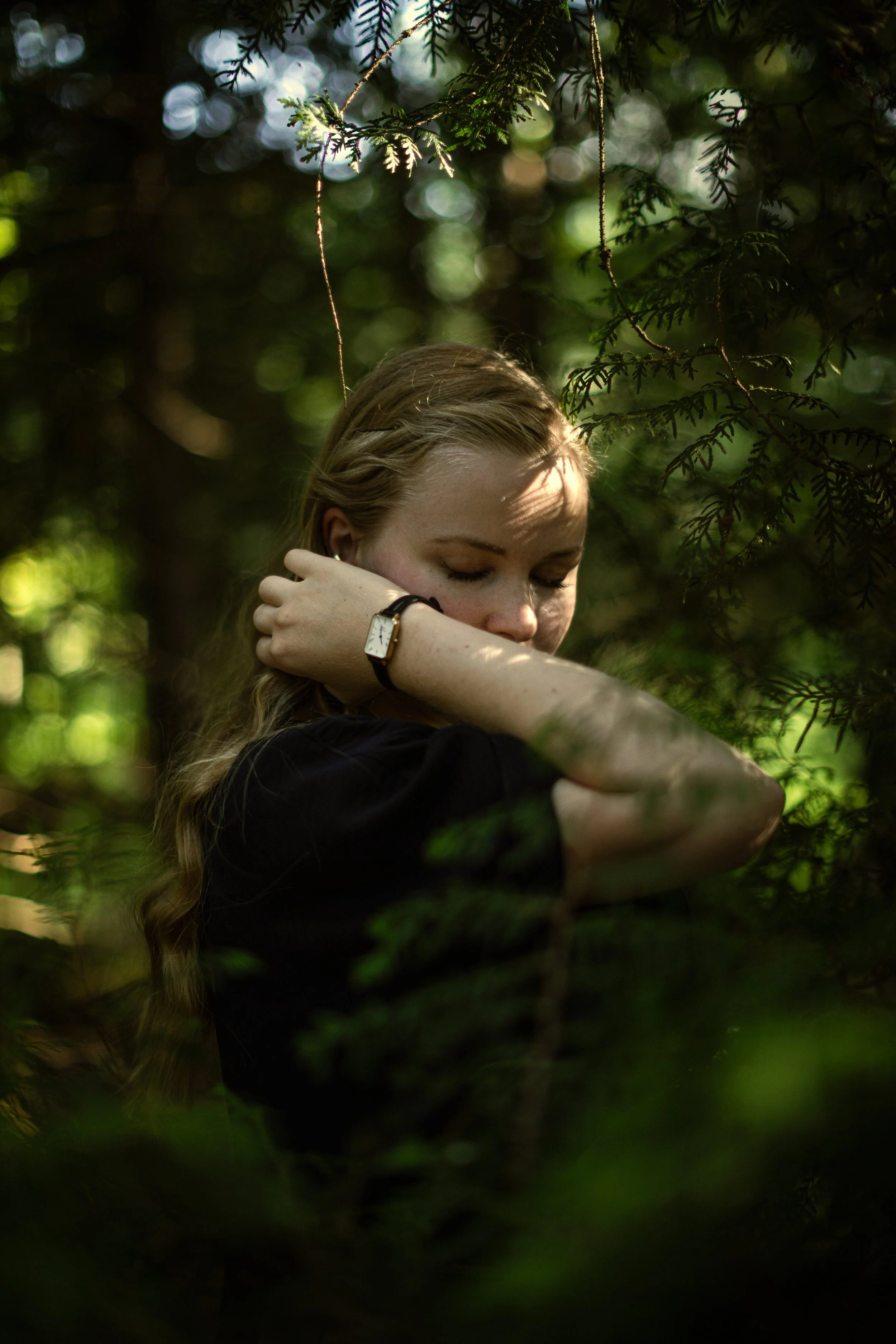 Junge Frau mit geschlossen Augen, Arm vor das Gesicht, steht im Grünen, trägt eine Uhr, im Schatten der Bäume.