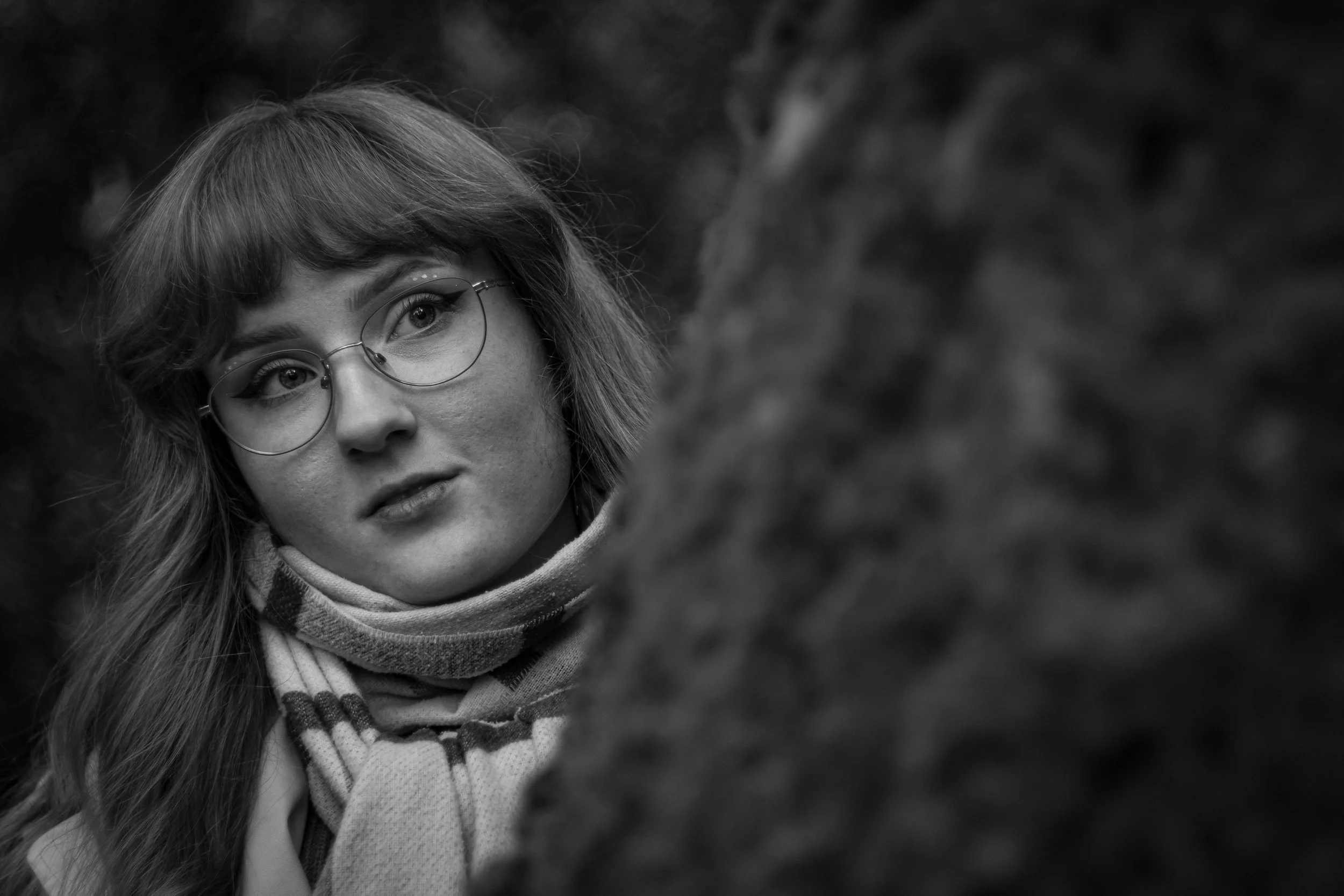 Nahaufnahme eines jungen Mädchens mit Brille und lockigem Haar, das nach links schaut, im Freien mit unscharfem dunklem Hintergrund.