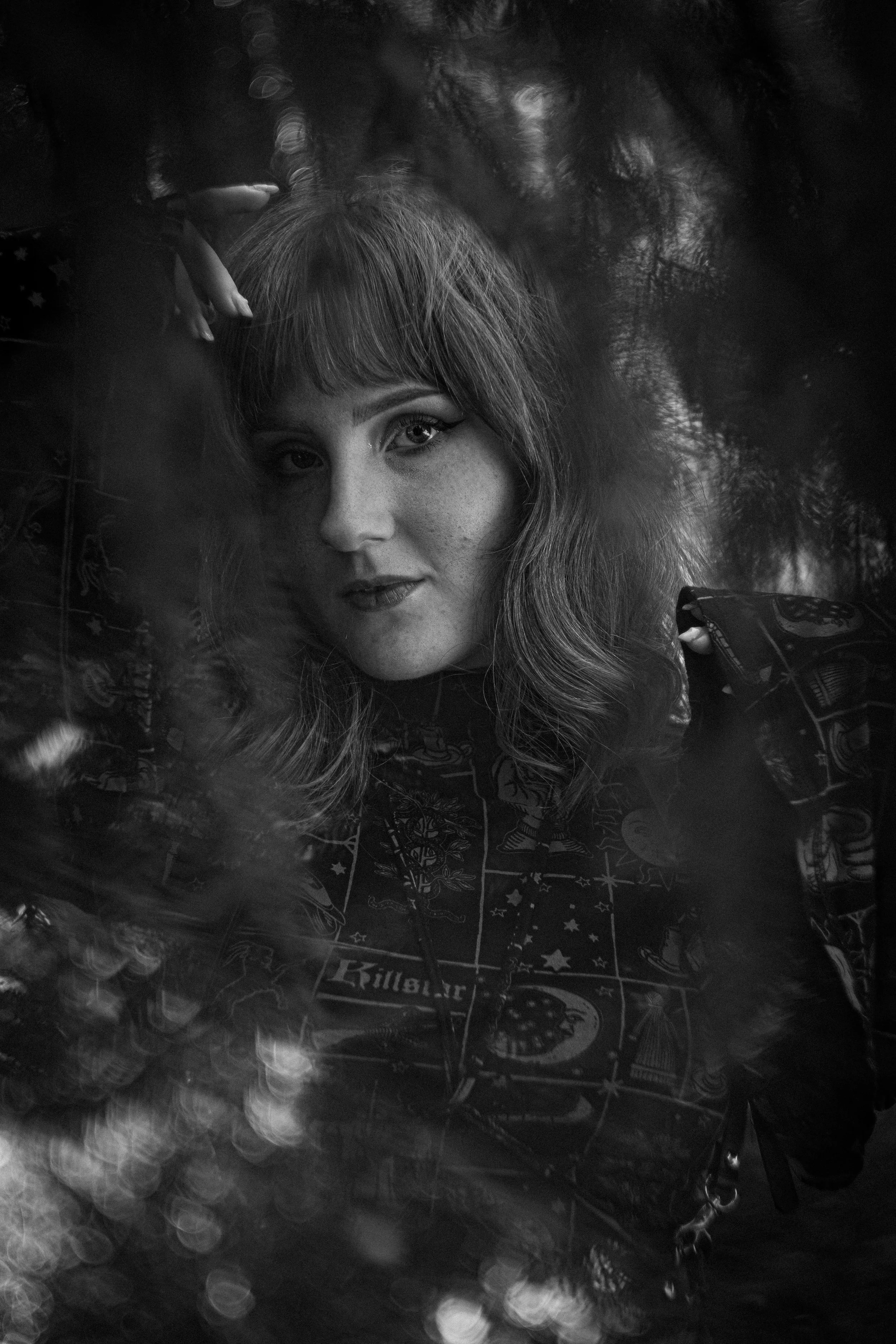 Schwarzweißfoto eines Mädchen mit langen, lockigen Haaren, das in eine Kamera schaut, teilweise deckend durch reflektierende Glasscheibe, im Hintergrund ein Tannenbaum