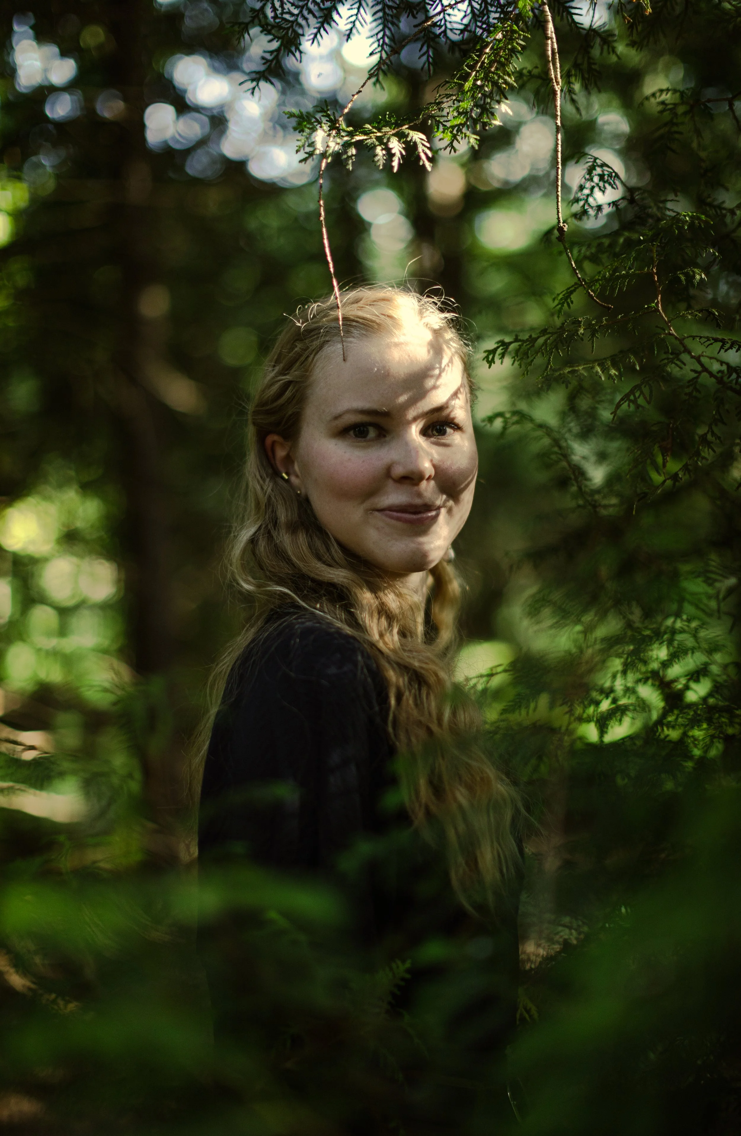 Junge Frau mit langen, welligen Haaren in einem dunklen Oberteil, die sich vor einem Hintergrund aus grünen Blättern und Tannenzweigen befindet. Das Licht fällt durch die Blätter und erzeugt einen Bokeh-Effekt.