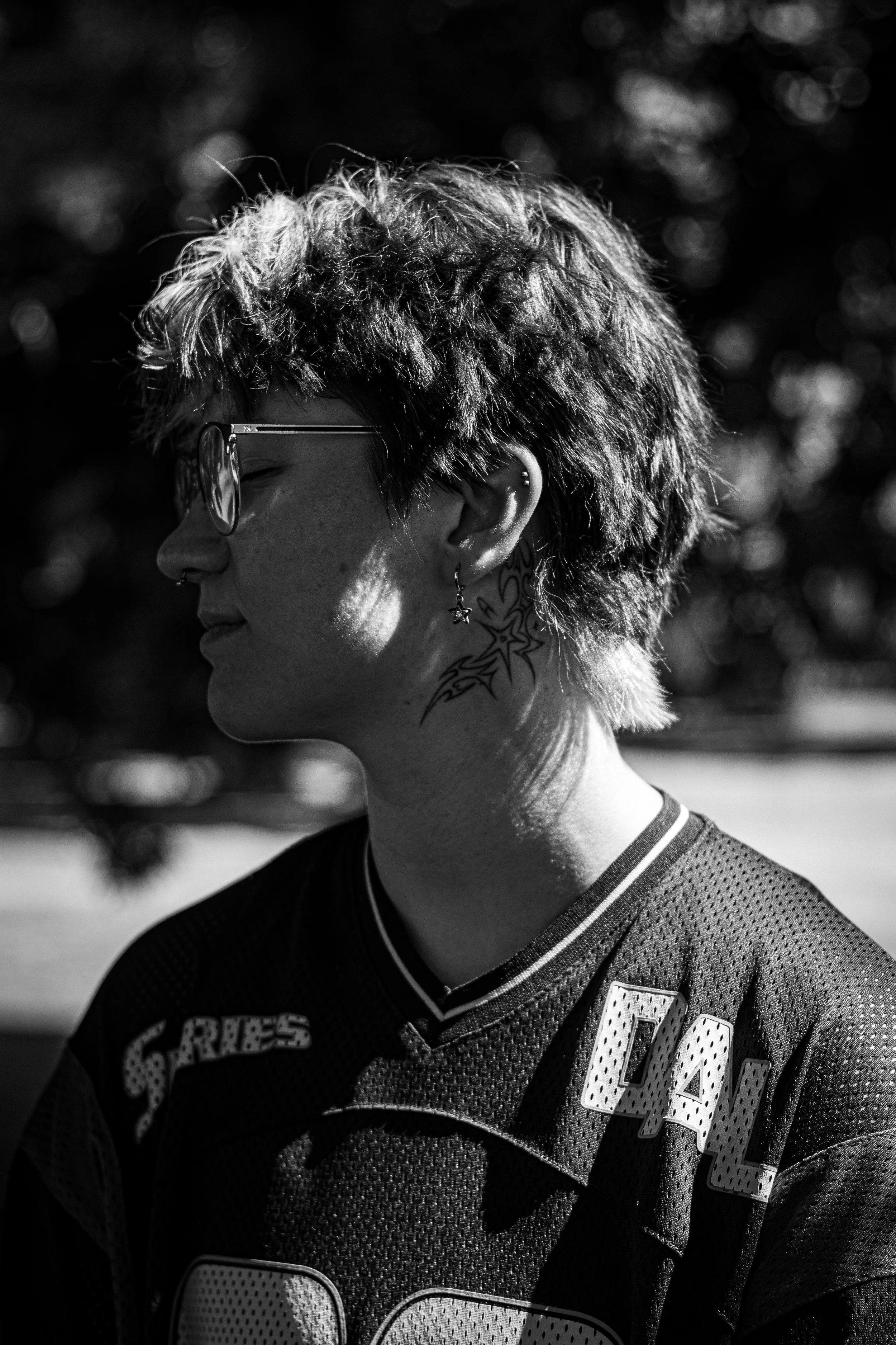 Nahaufnahme eines jungen Mannes mit kurzen, lockigen Haaren, Brille, Ohrring und Tattoos am Hals, trägt ein trikotartige Kleidung, im Profil, im Sonnenlicht, unscharfer Hintergrund.