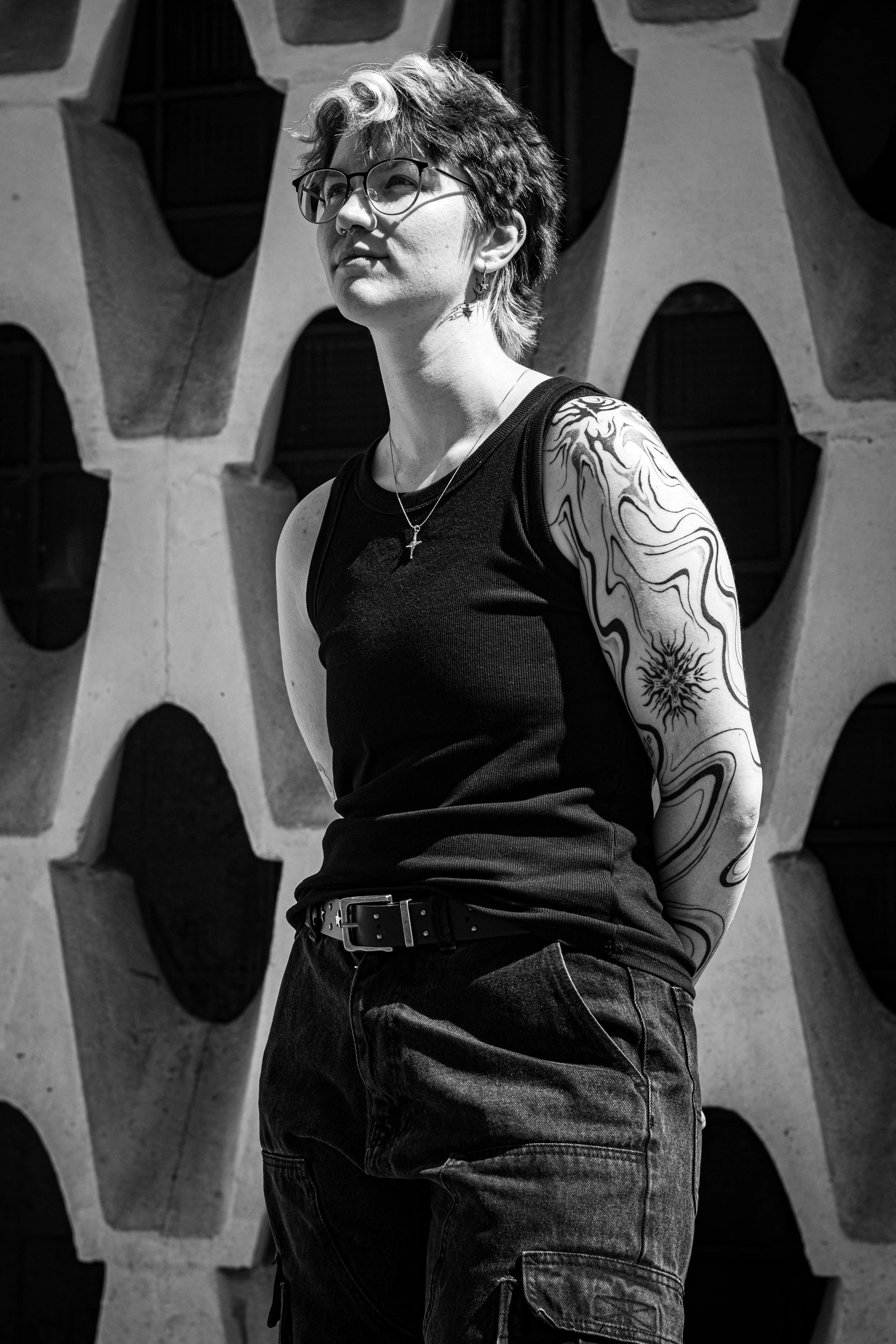 Schwarz-weißer Foto einer jungen Frau mit Brille, Tattoos am Arm, kurzen Haaren und einem Kreuzhalskette, stehend vor einer Architektur aus Stein mit geometrischen Mustern.
